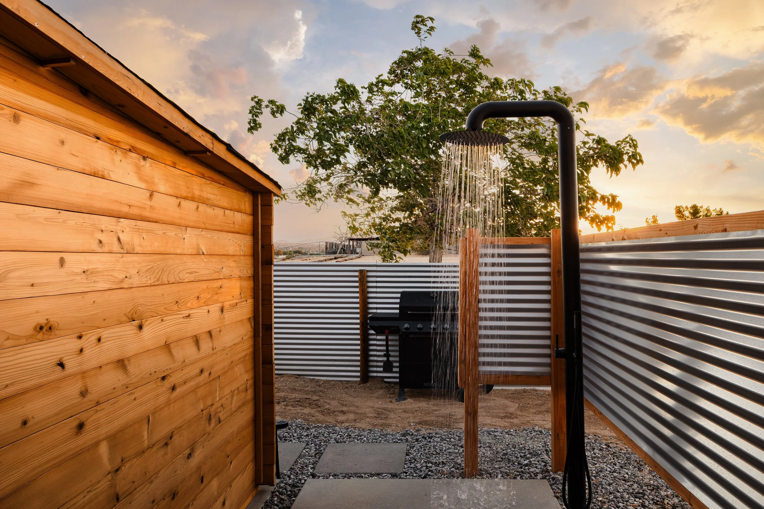 An outdoor shower with a rain shower head mounted on a black pipe, next to a wooden fence and a metal fence, with a tree and cloudy sky in the background, during sunset.