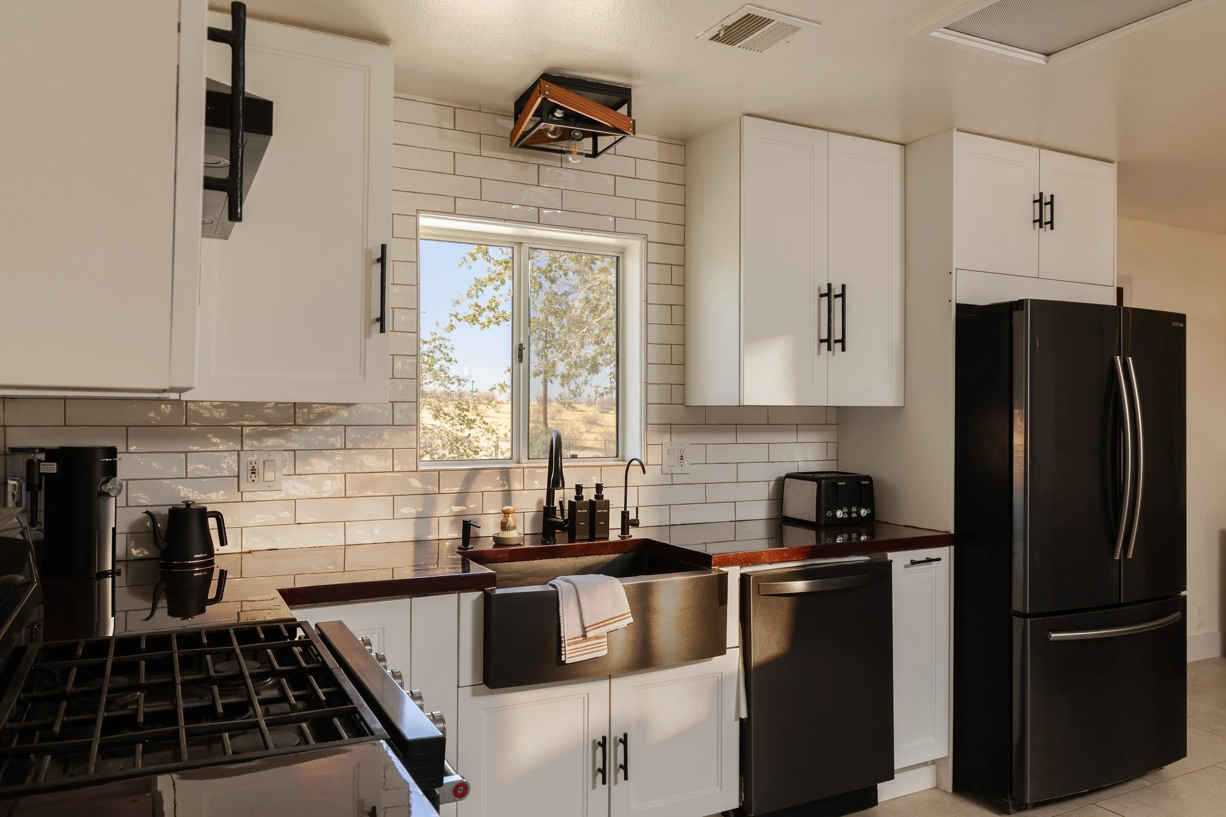 Modern kitchen with white cabinets, black handles, a window above the sink, and appliances including a black refrigerator, stove, and small black toaster. There is a white tiled backsplash and a ceiling light fixture.