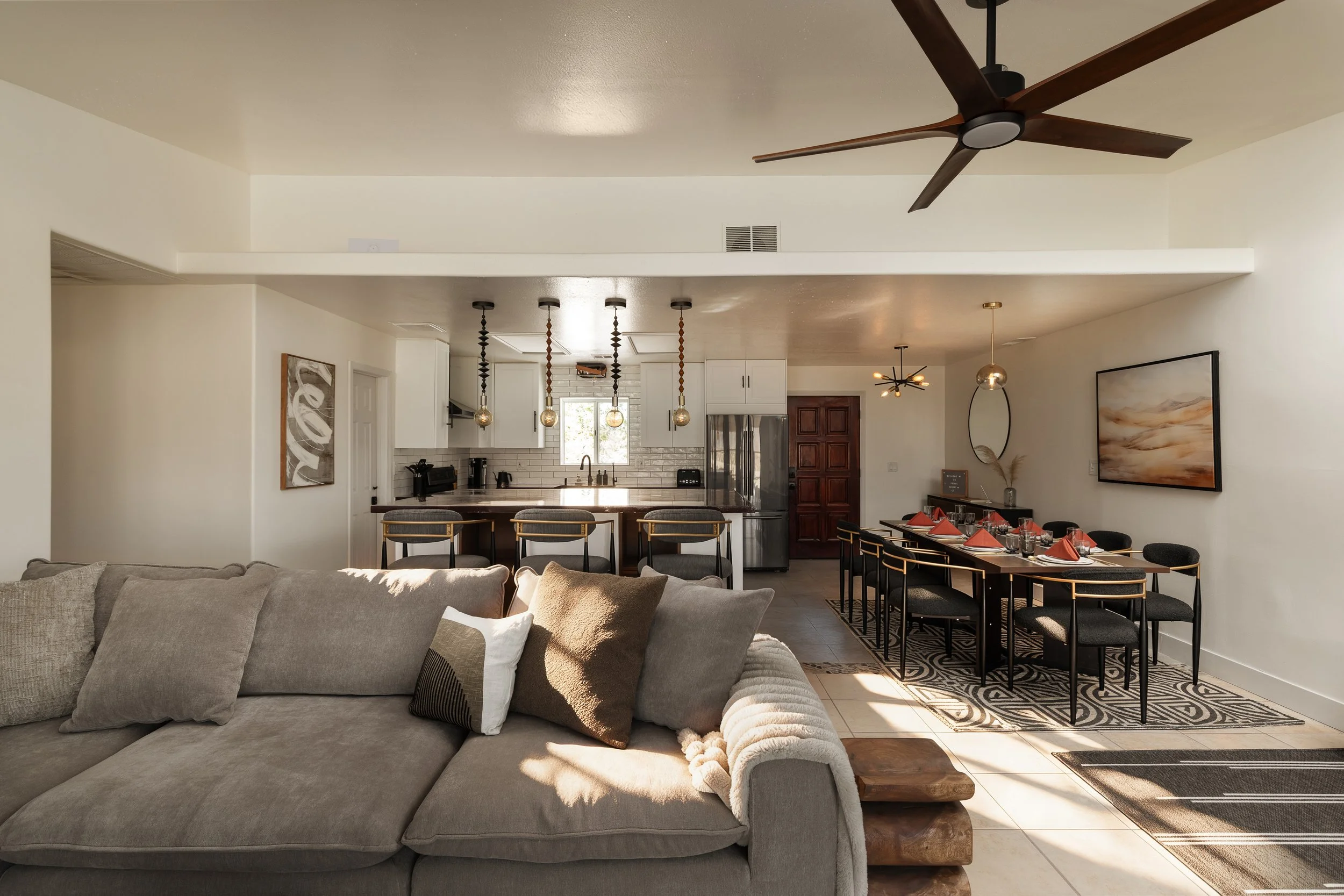 Open-concept living space with a beige sectional sofa in the foreground, and a dining area with a black table and chairs set with red napkins, and a kitchen with white cabinets and pendant lights hanging over a kitchen island in the background.