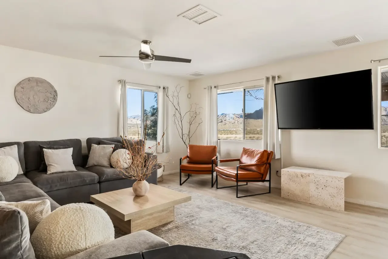Living room with a gray sectional sofa, white pillows, two orange chairs, a coffee table with a vase, a wall-mounted flat-screen TV, windows with white curtains, and mountains visible outside.