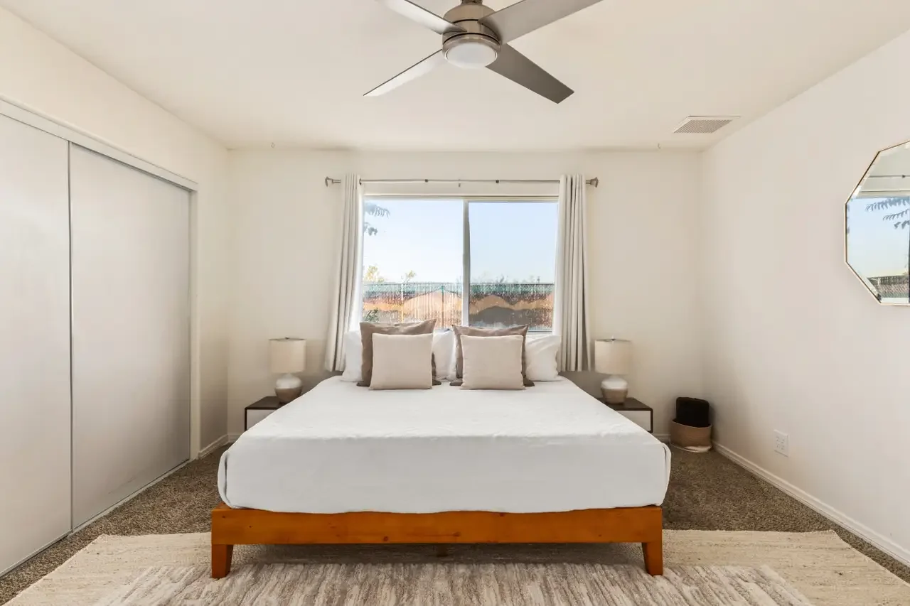 A bedroom with a bed featuring white bedding and pillows, two bedside tables with lamps, a large window with white curtains, a ceiling fan, and a mirror on the wall.