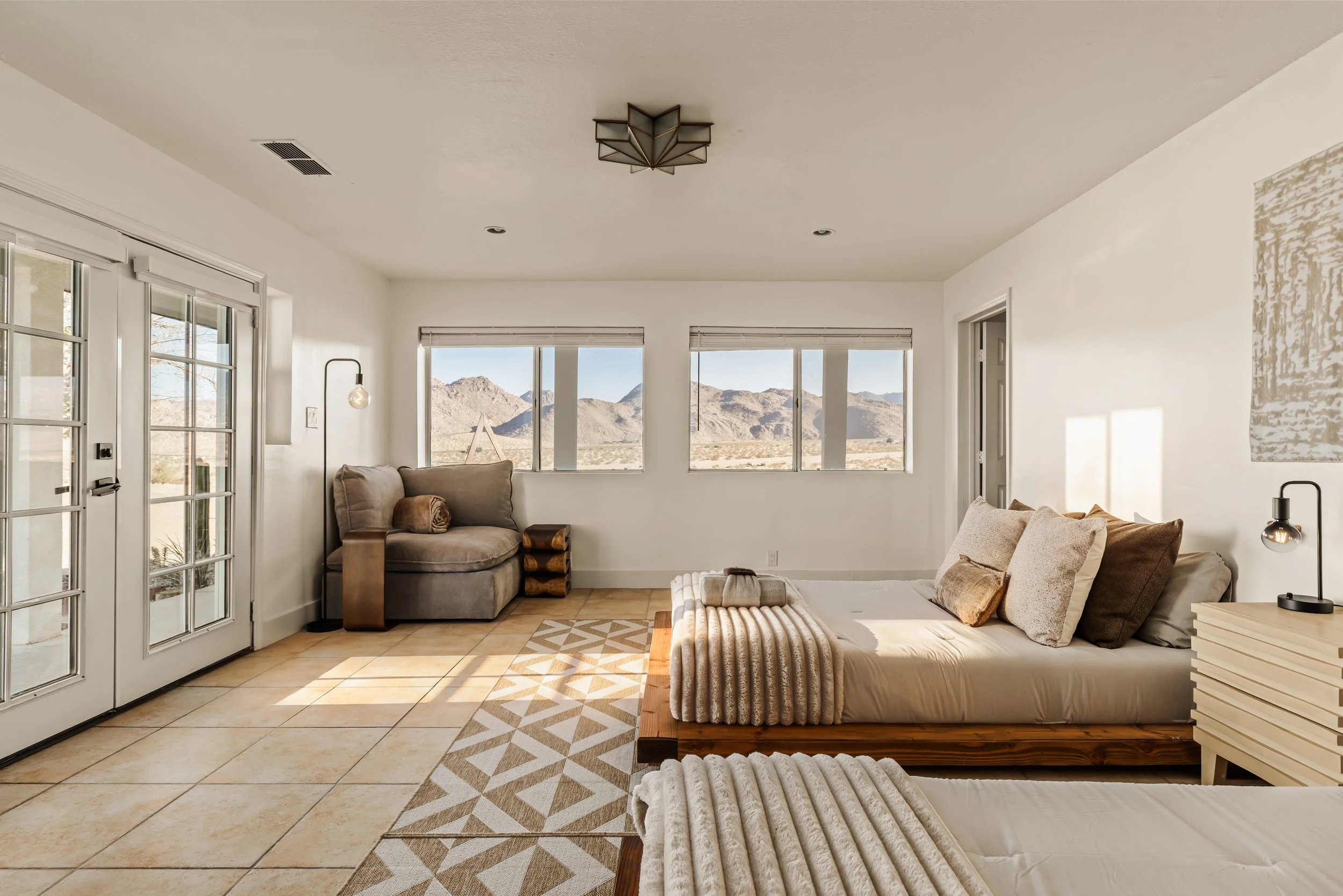 A bright living room with beige tiled floor, white walls, and large windows showing mountainous desert landscape. There is a beige sofa with brown and cream cushions, a wooden slatted bench, and a patterned area rug. French doors lead outside, and th