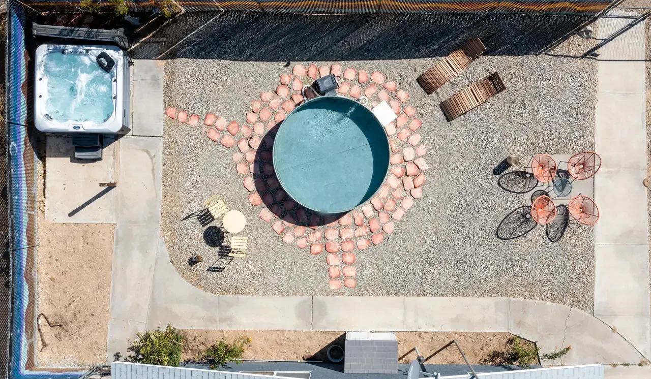 A backyard with a hot tub in the upper left corner, a small above-ground pool in the center surrounded by pink stones, four orange chairs on the right side, two brown benches at the top, a small table with four chairs near the pool, and a concrete si