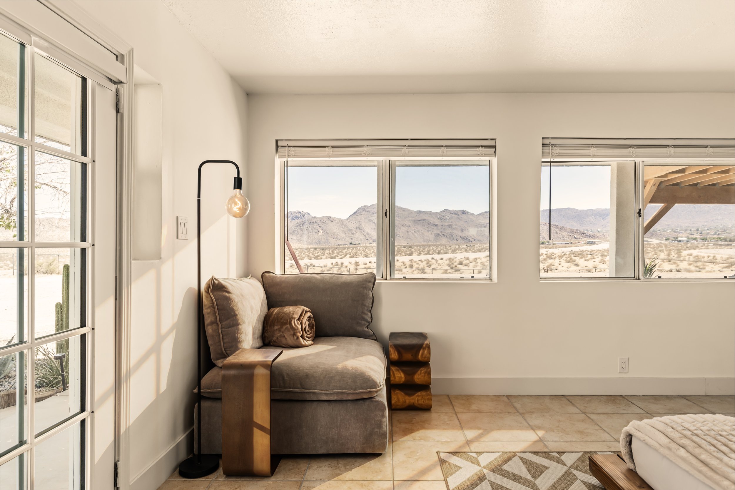 A cozy living room with a beige armchair, a side table, a wall-mounted lamp, and large windows offering a mountain view in the desert.