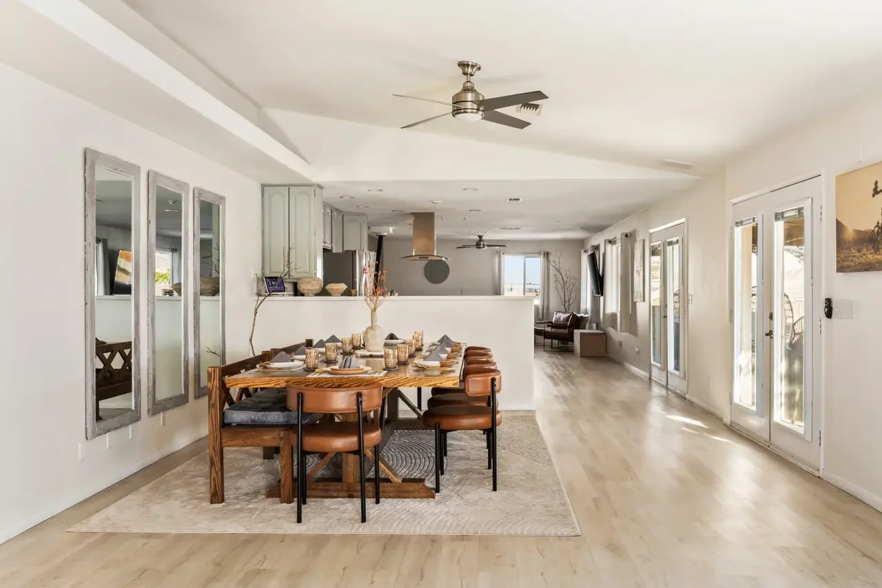 Open-concept dining and living space with a wooden dining table set for six, beige chairs, wall mirrors, white walls, and multiple glass doors leading outside. Kitchen in background with stainless steel appliances and a ceiling fan.