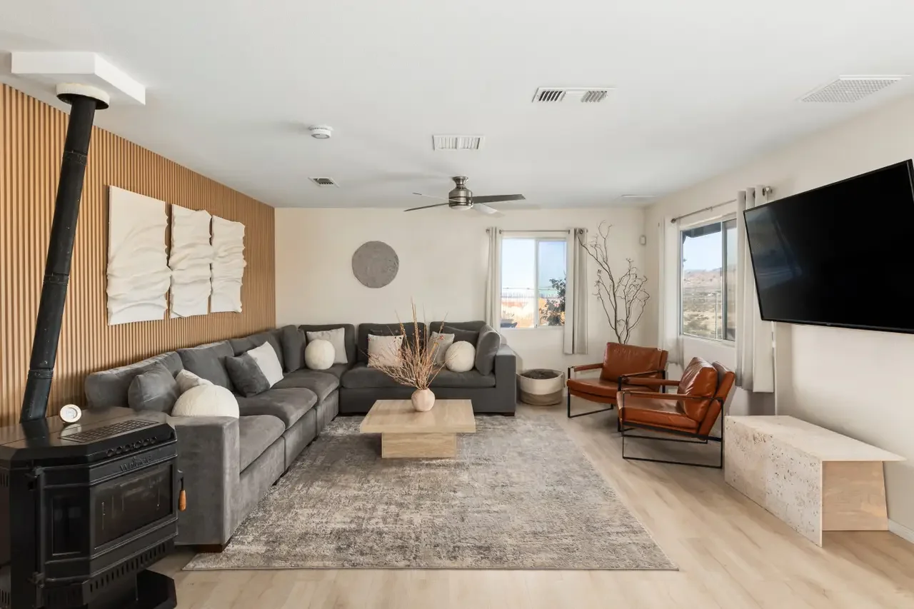Living room with a gray sectional sofa, two brown leather chairs, a wooden coffee table, a wall-mounted TV, and large windows with white curtains.