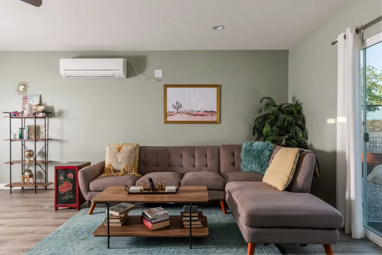 Living room with beige sectional sofa, coffee table, wall art, bookshelf, potted plant, and sliding glass door with curtains.