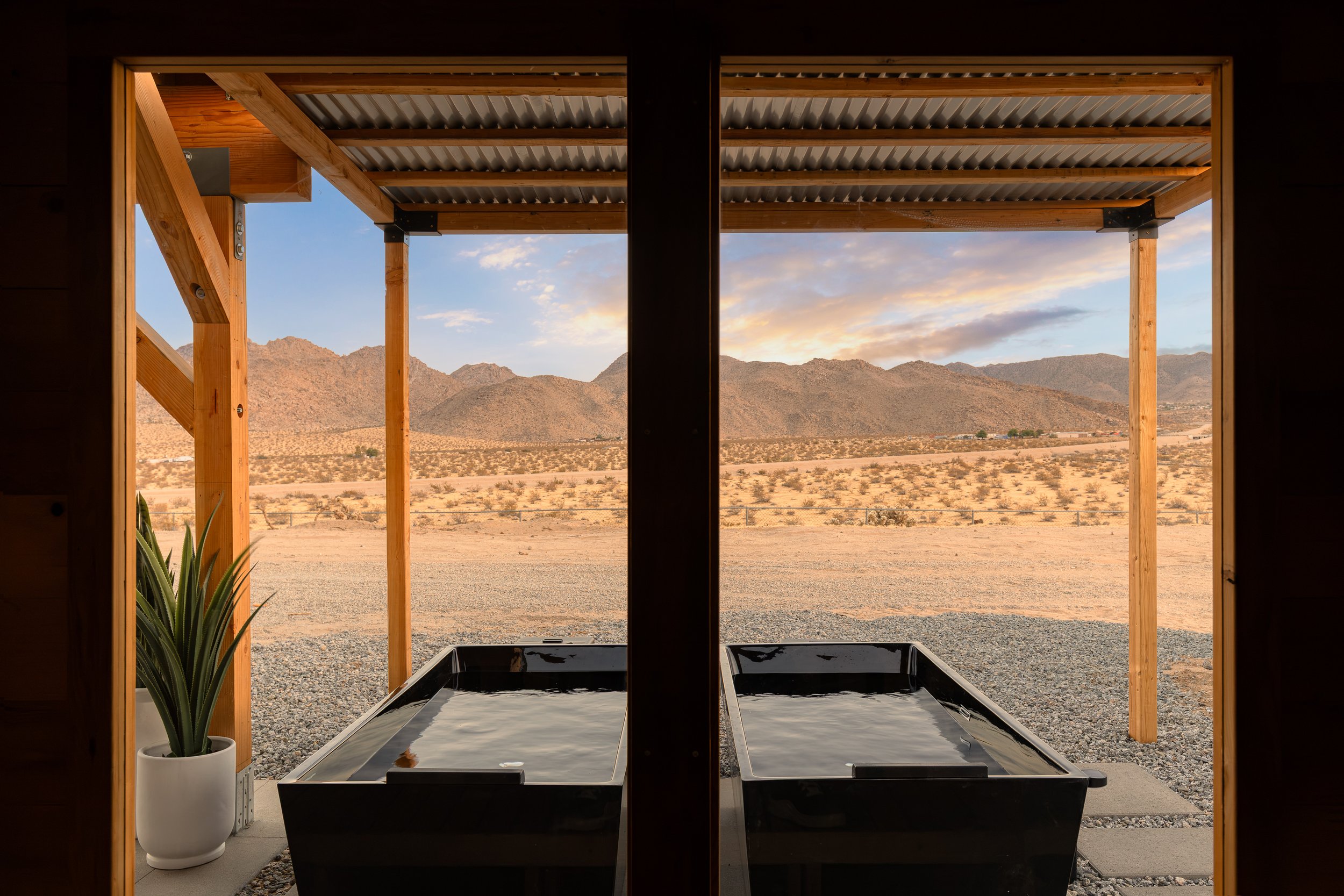 View through a doorway showing an outdoor desert landscape with mountains in the distance, a wooden structure with a corrugated metal roof, and two black soaking tubs.