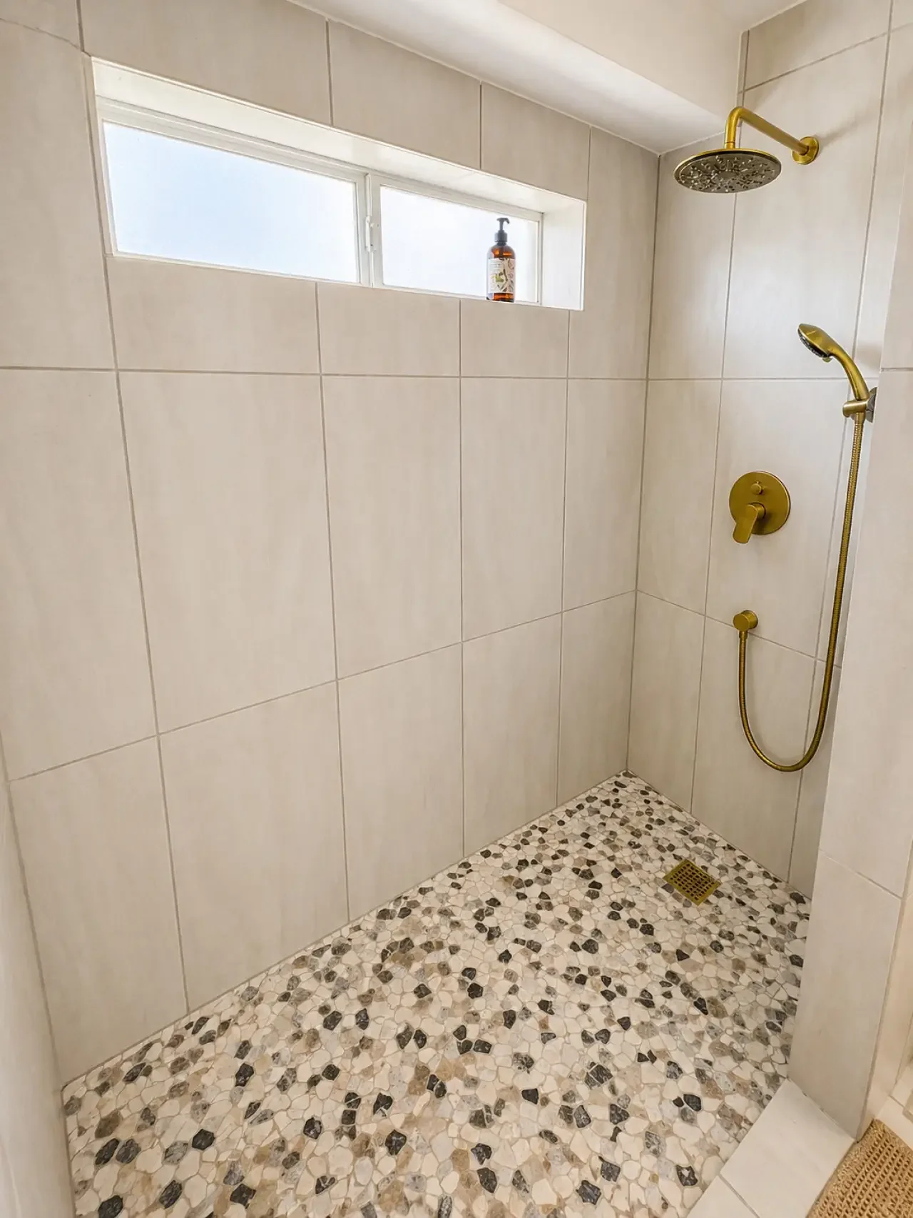 A walk-in shower with beige tiles, a mosaic pebble floor, a gold showerhead, a handheld shower, a small high window with frosted glass, a bottle of soap or shampoo on the windowsill, and a floor drain.