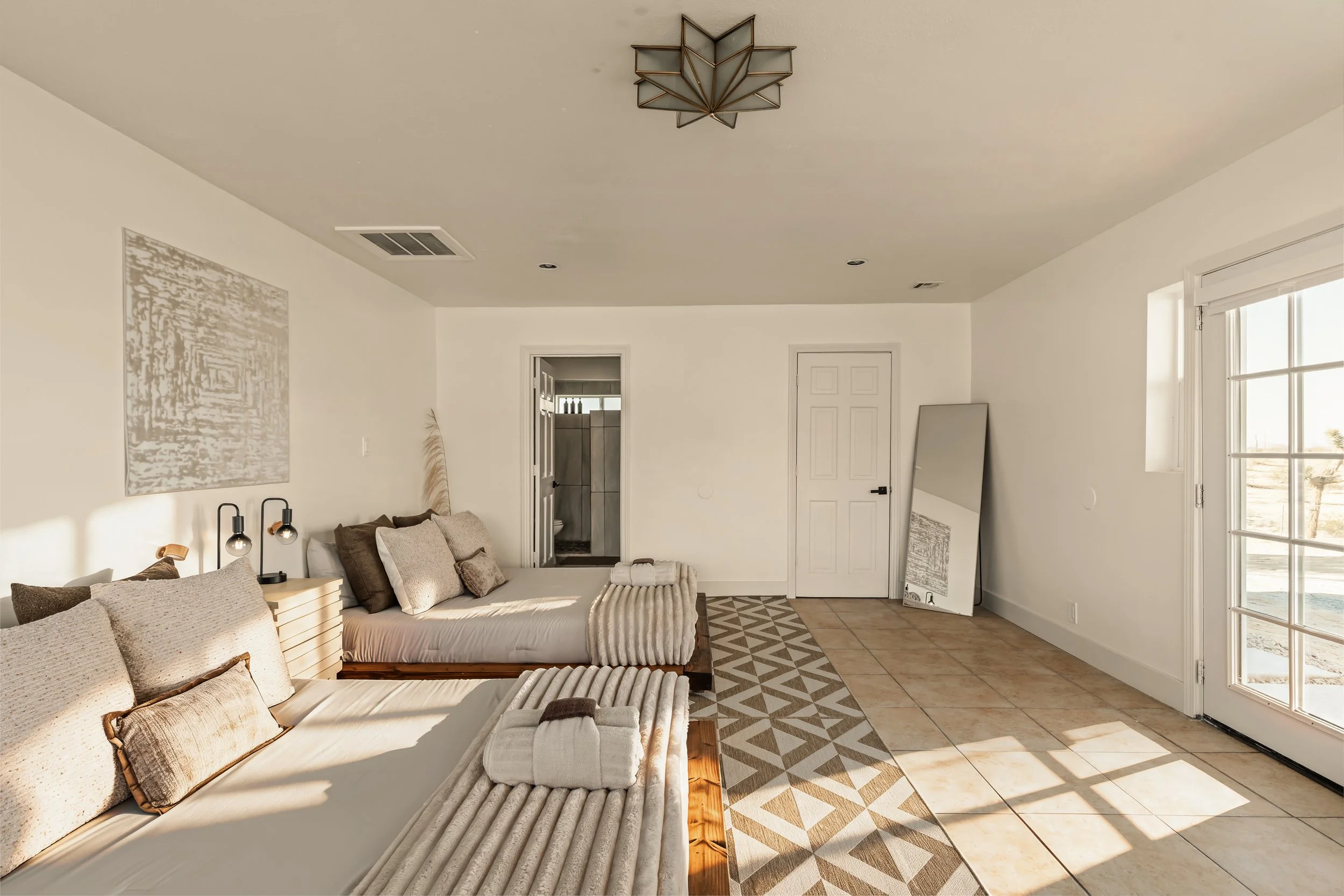 Living room with beige couches, pillows, a patterned rug, and a large window letting in sunlight, with a door leading to a bathroom.