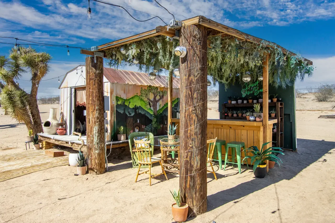 A small outdoor desert bar with a wooden structure, string lights, potted plants, and seating, in a sandy area under a blue sky.