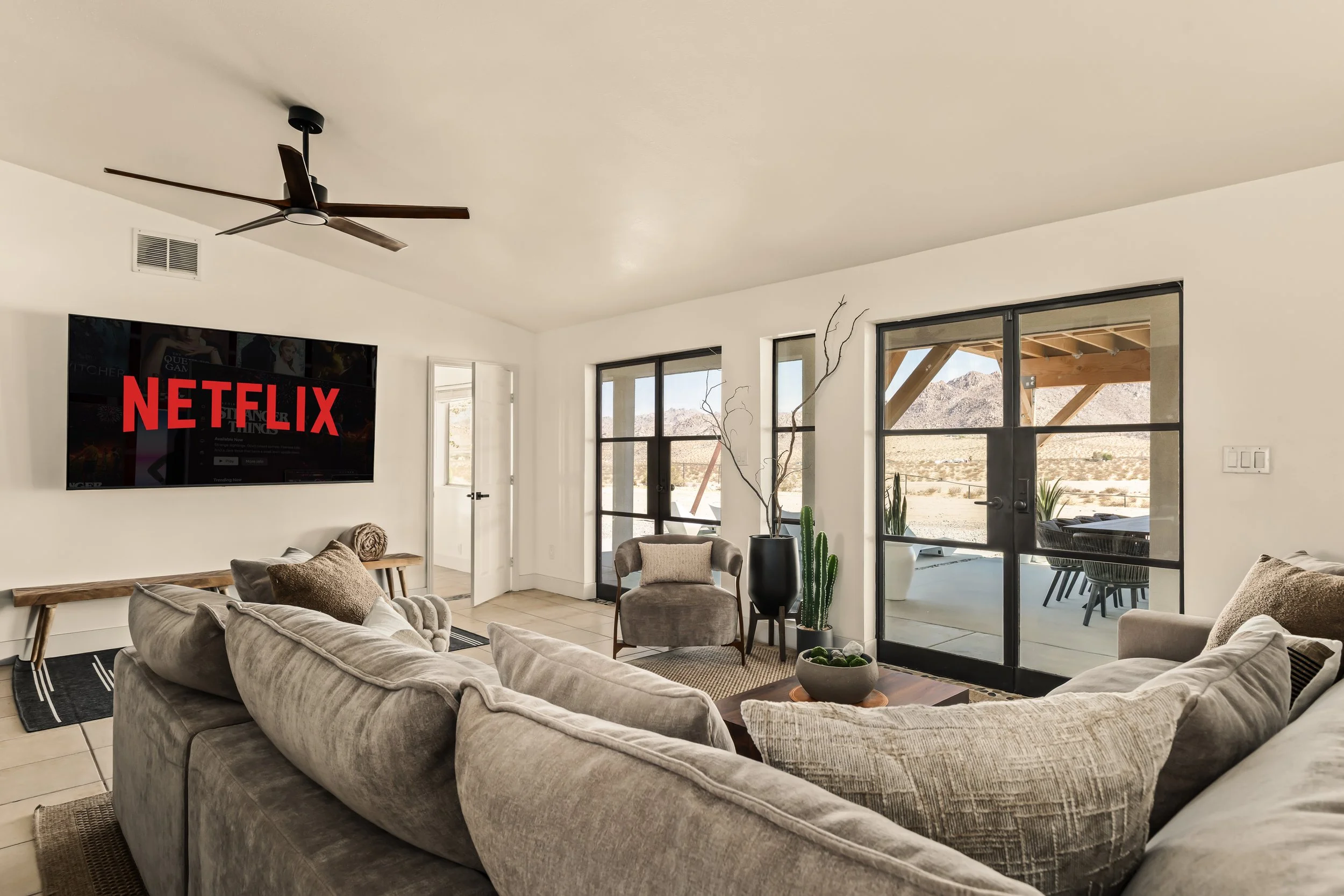 Living room with beige couches, a wall-mounted Netflix TV, large windows with mountain views, a black sliding door, and desert landscape outdoors.