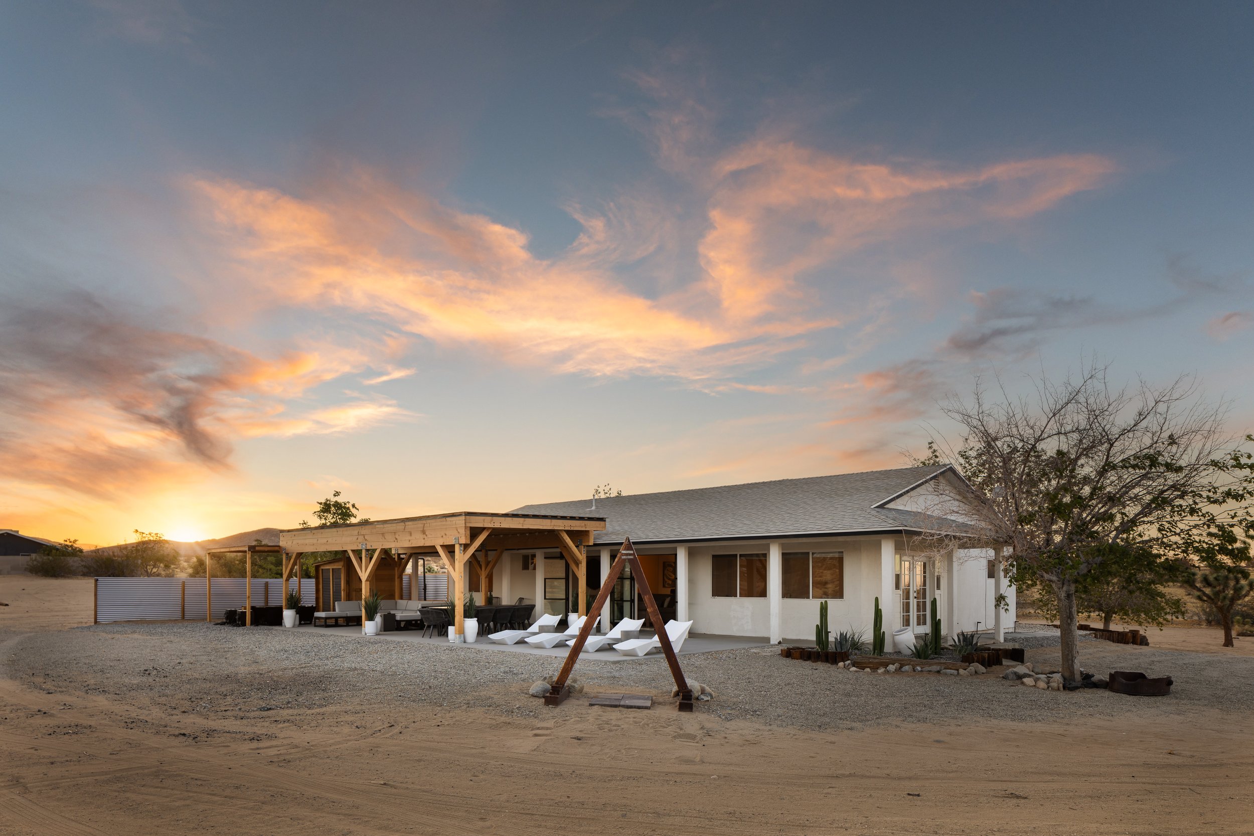 A modern white house with a gray roof, outdoor seating area, and swing set in the backyard during sunset, with a desert landscape and mountains in the background.