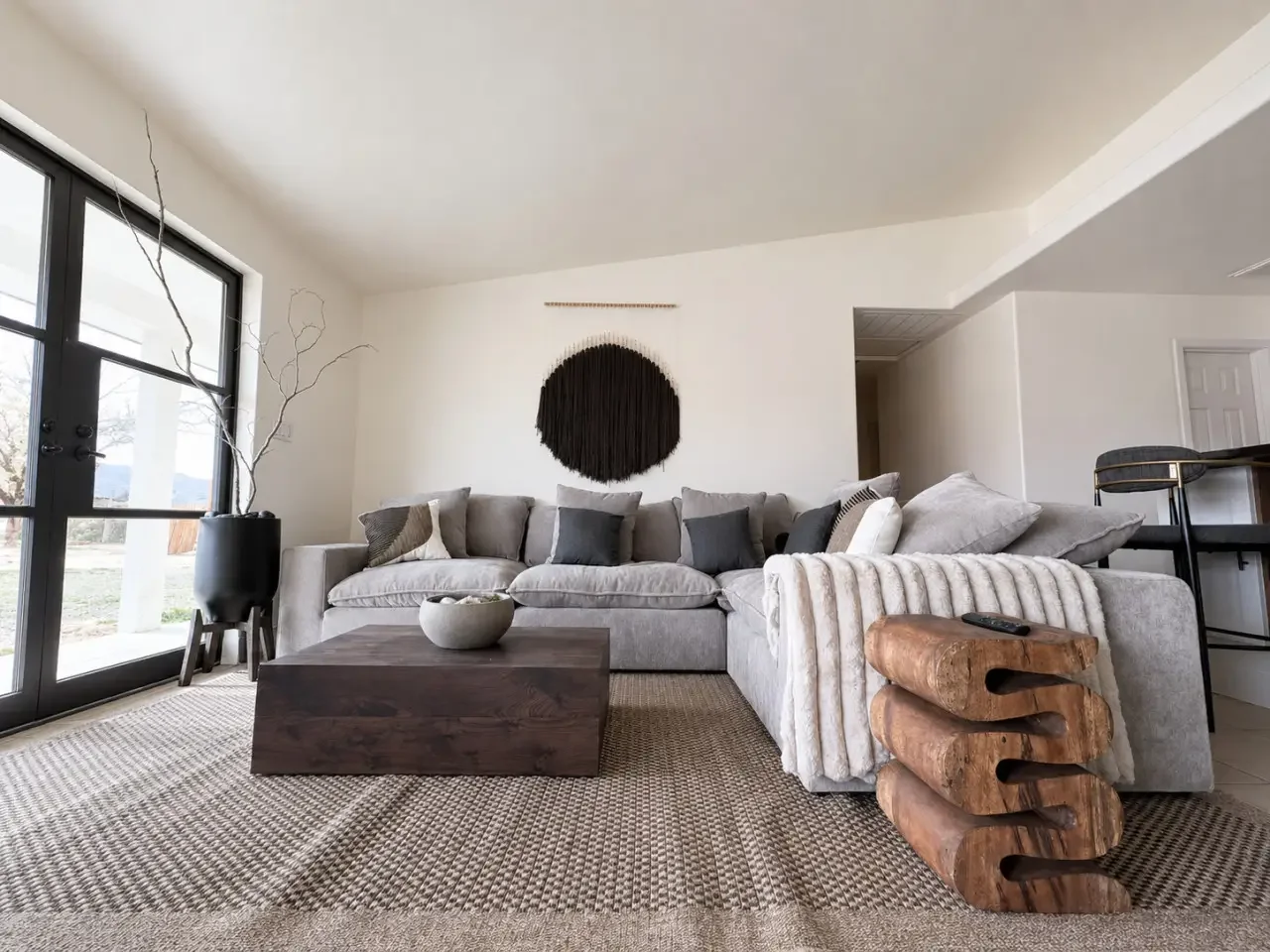Living room with a large gray sectional sofa, dark throw pillows, a wooden coffee table, a vase with bare branches, and decorative wooden logs stacked on the side, with a large round black wall hanging and a window letting in natural light.