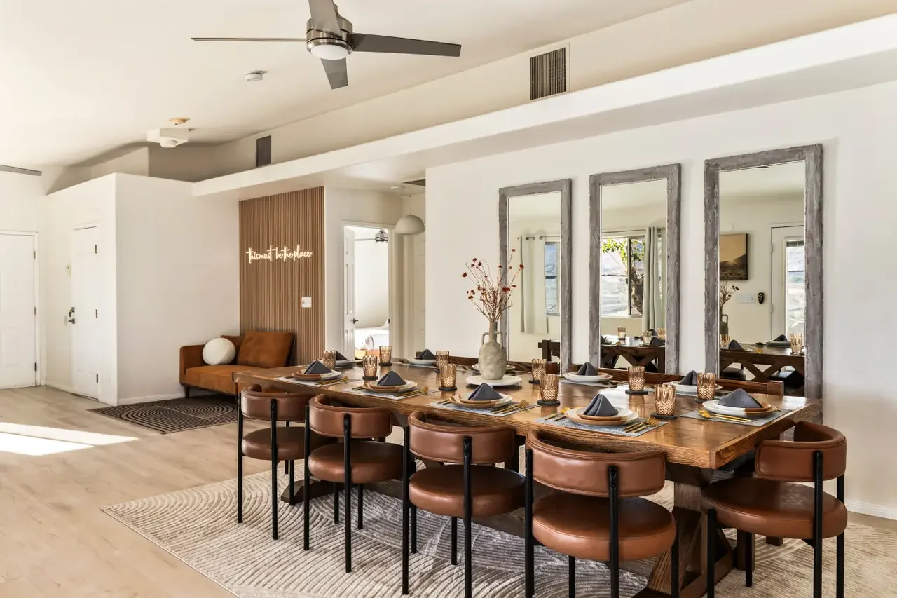 Dining room with a large wooden table set for a meal, surrounded by brown chairs, and three tall mirrors hanging on the wall behind it. A decorative vase with dried flowers is placed on the table. The room has natural light coming from the windows, a