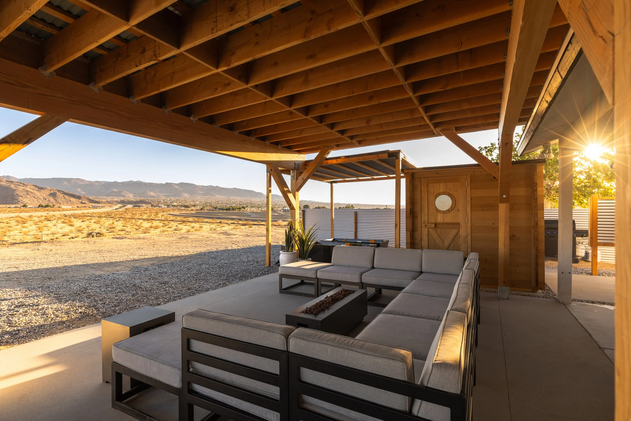 An outdoor patio with modern gray sectional sofa, fire pit table, and wooden shelter, overlooking a desert landscape with distant mountains and the setting sun.