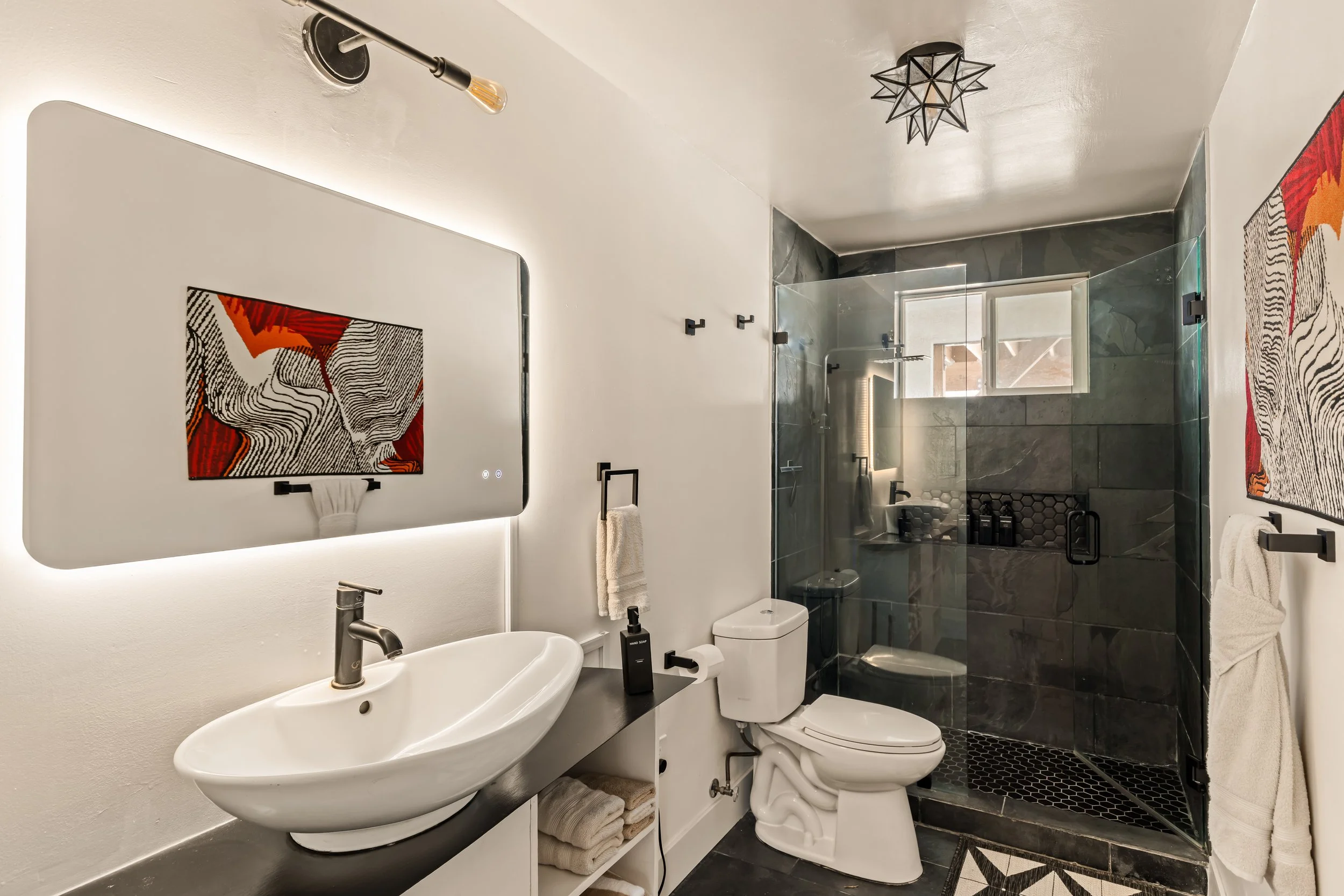 Modern bathroom with a white sink, black countertop, and a large mirror. The shower area has dark tile walls, a glass door, and a window. Artworks and towels are visible.