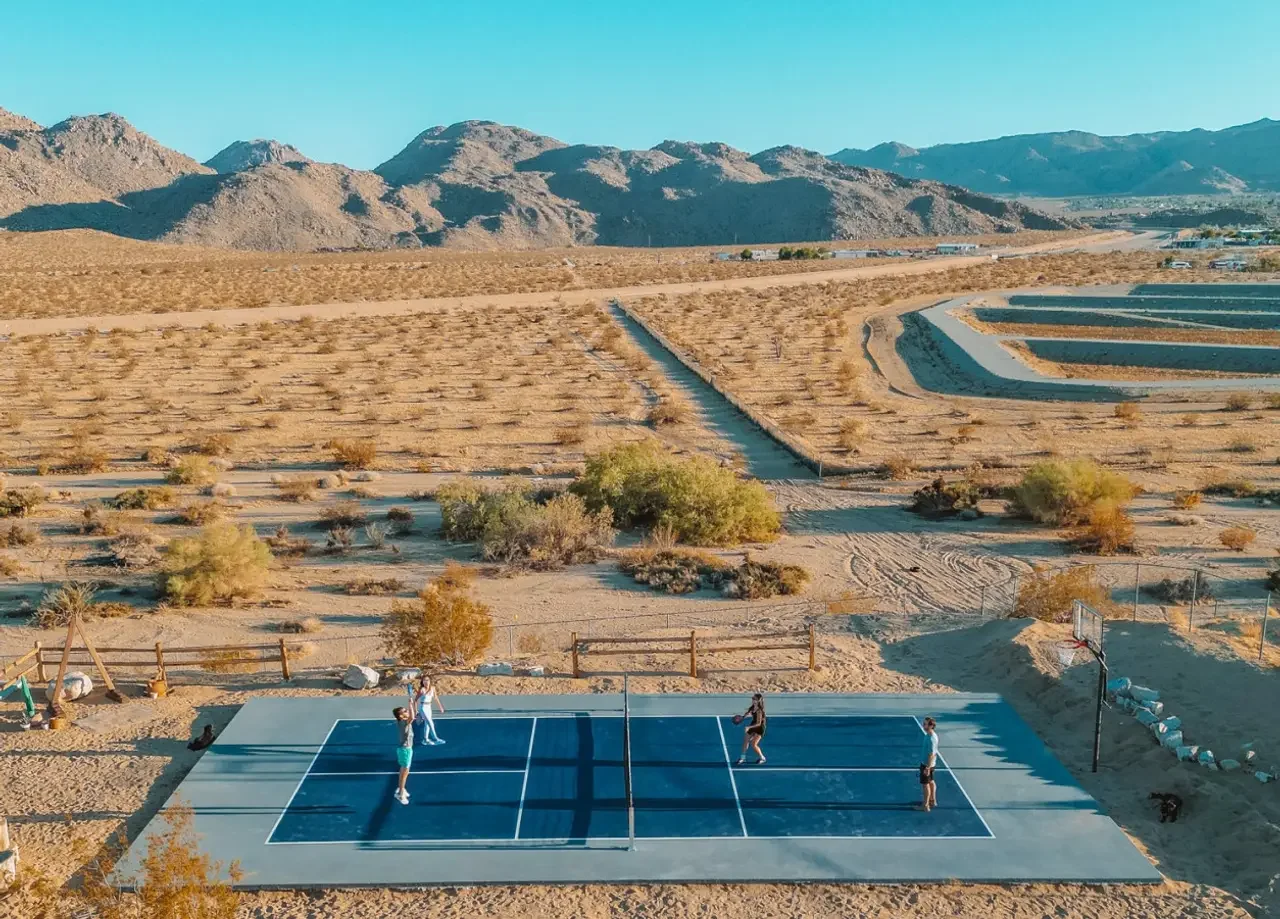 Four people playing pickleball on an outdoor court in a desert with mountains in the background, adjacent to a vineyard or orchard with rows of plants.
