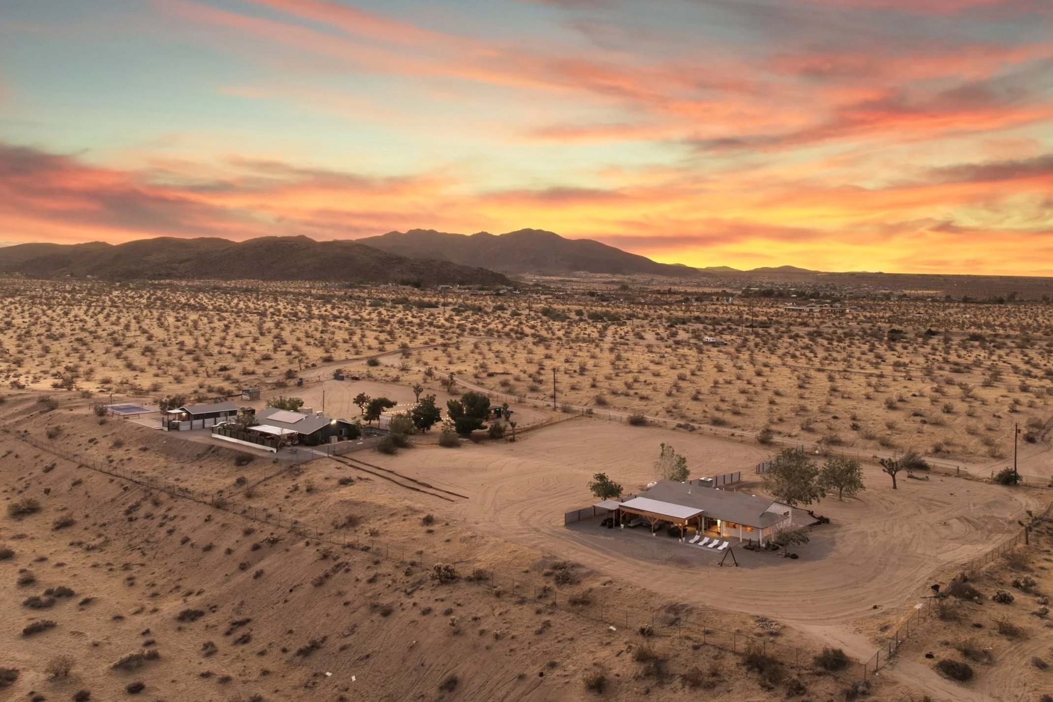 A desert landscape at dusk with colorful clouds in the sky, mountains in the distance, and a small residential area with houses and dirt roads in the foreground.