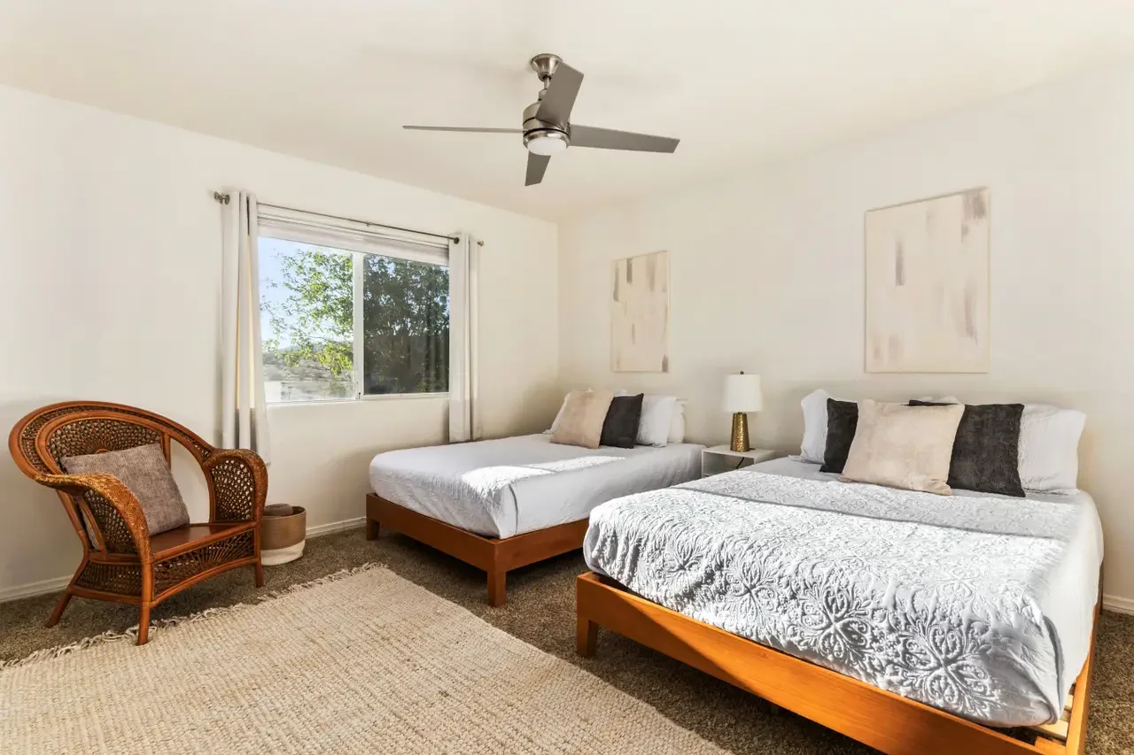 Bedroom with two queen-sized beds, a window with white curtains, a rattan chair with a cushion, a beige area rug, abstract wall art, a white nightstand, and a silver ceiling fan.
