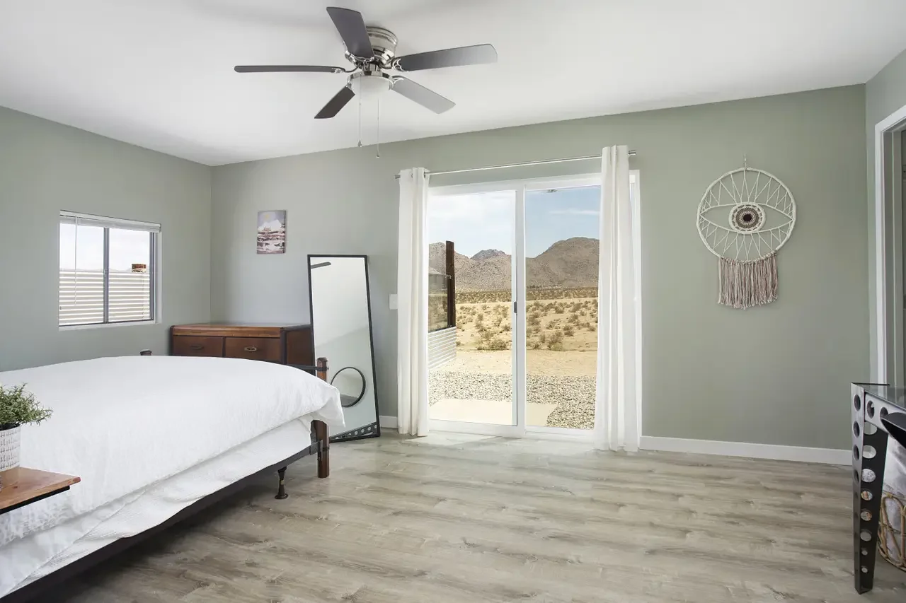 A bedroom with a bed, a small dresser, a floor mirror, and wall decor. Open sliding glass door shows desert landscape with mountains.