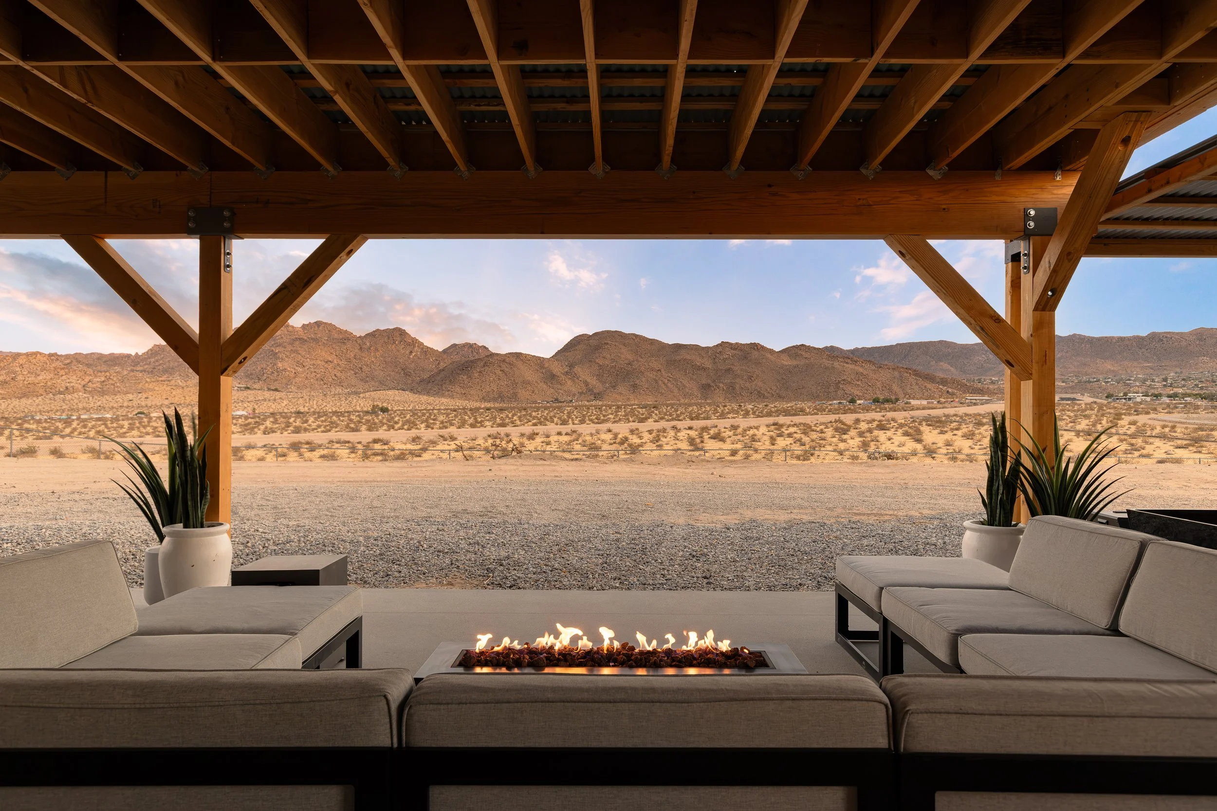 Outdoor seating area with couches and fire pit under a wooden pergola, overlooking desert mountains and sky.