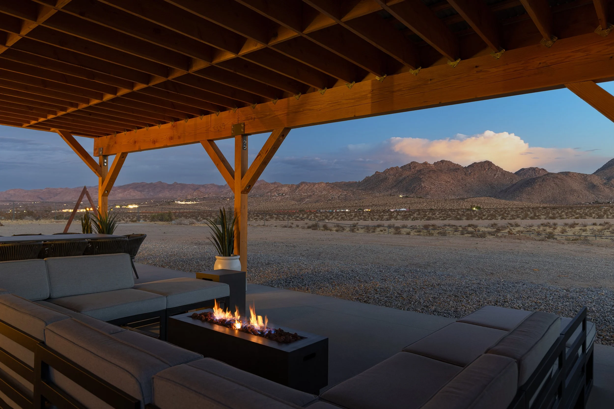 Outdoor seating area with a fire pit, patio furniture, a wooden overhead canopy, and desert mountains in the background during sunset.