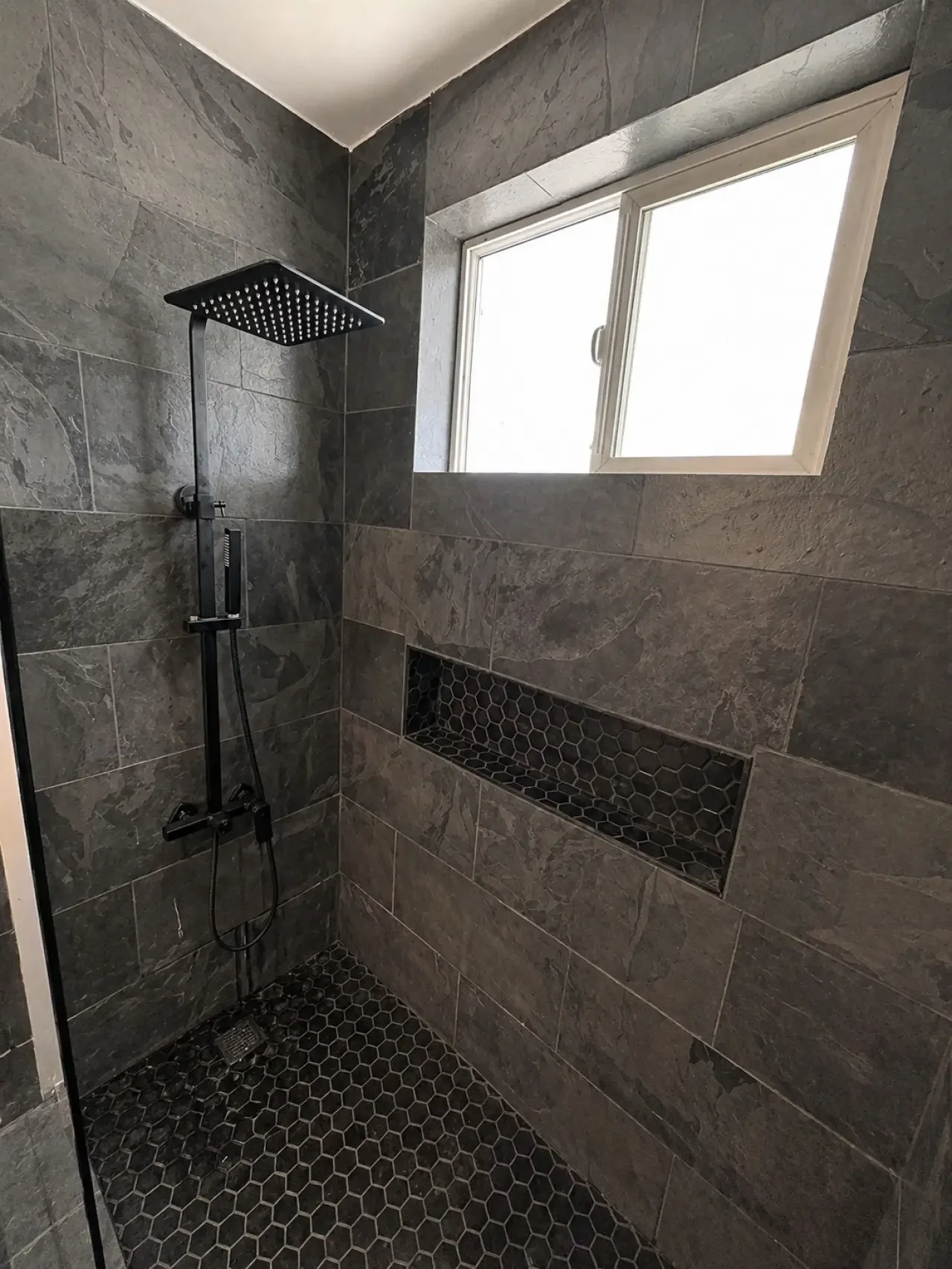 A modern shower with dark gray stone wall tiles, a black rain showerhead, a built-in niche with a black hexagon tile pattern, and a window with frosted glass.
