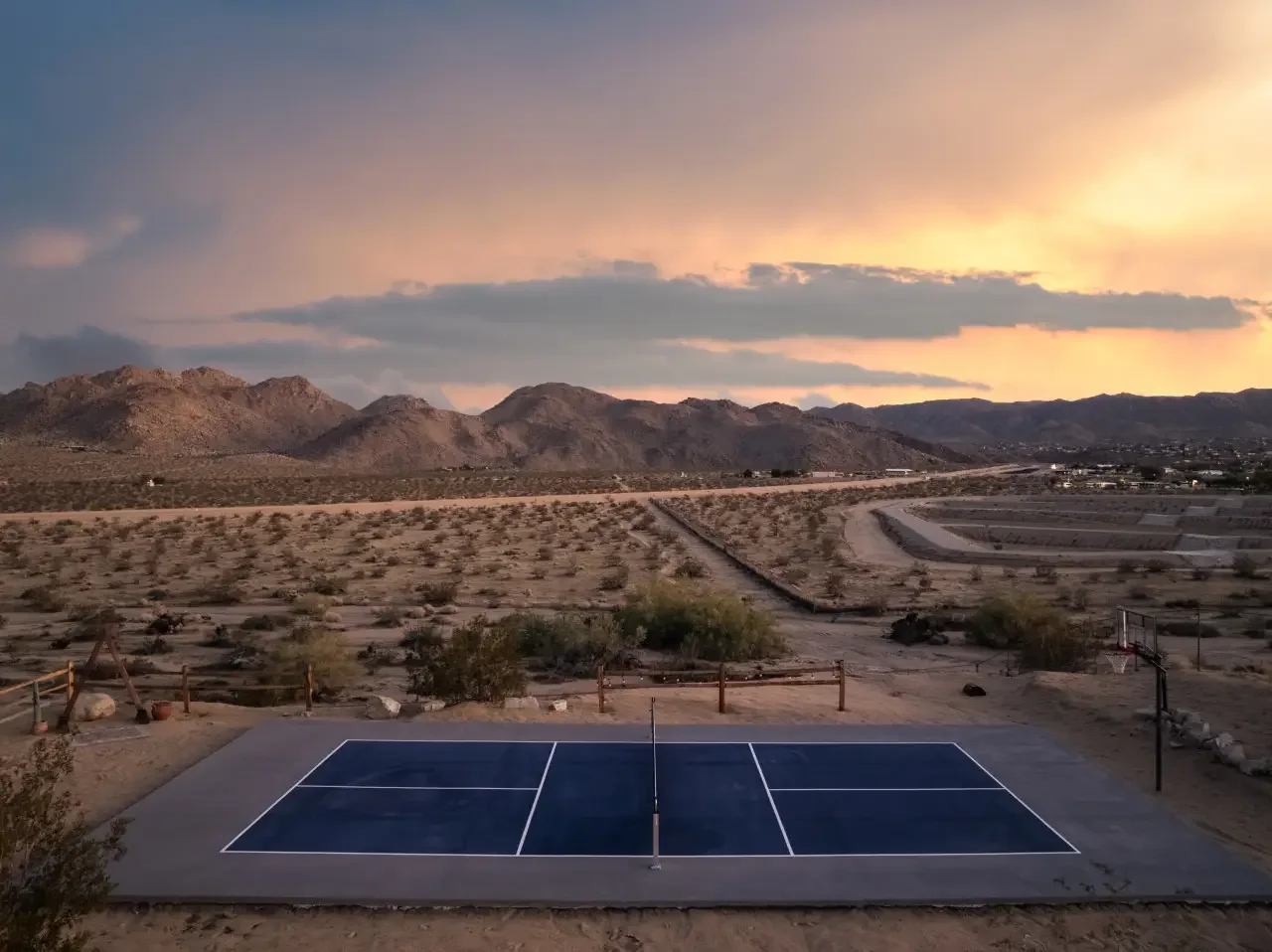 A blue outdoor tennis court in a desert landscape at sunset with mountains in the background and a cloudy sky.