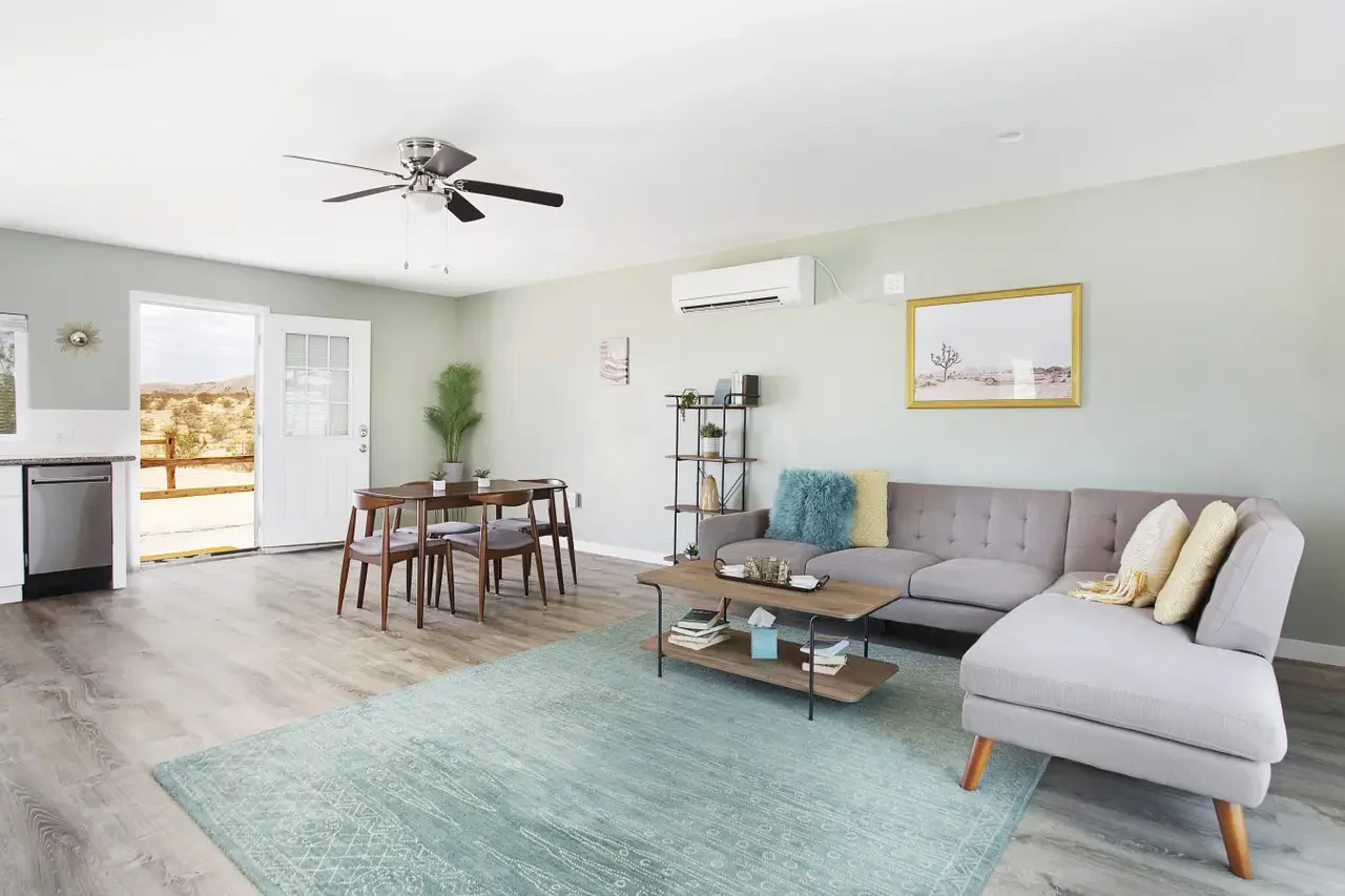 Living room with gray sectional sofa, wooden coffee table, teal and yellow pillows, green rug, dining table with wooden chairs, potted plants, and a door leading outside, with a ceiling fan and an air conditioning unit on the wall.
