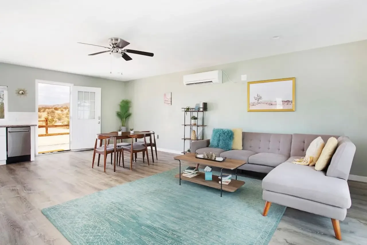 Bright living room with a gray sectional sofa, teal and yellow pillows, a wooden coffee table, a teal area rug, a dining table with chairs near a door leading outside, a black shelving unit with decor, a tall green plant, a wall photo, an air conditi