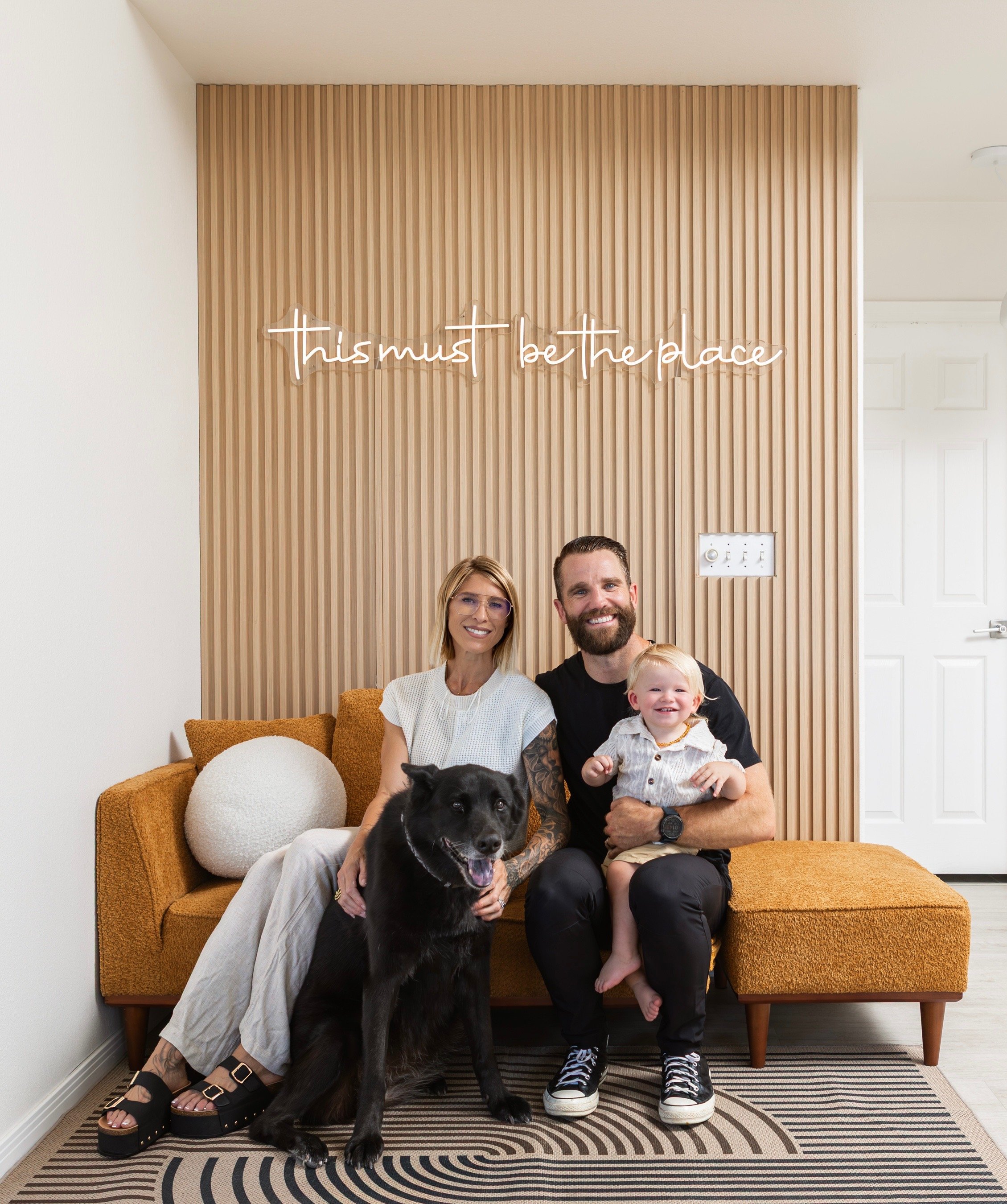 A smiling family of three with a dog, sitting on a mustard-colored sofa in a modern living room. The woman has blonde hair and glasses, the man has a beard, and the child is a toddler with light hair. A neon sign above them reads 'this must be the place.' The room features a wooden slat accent wall, a white door, and a patterned rug.