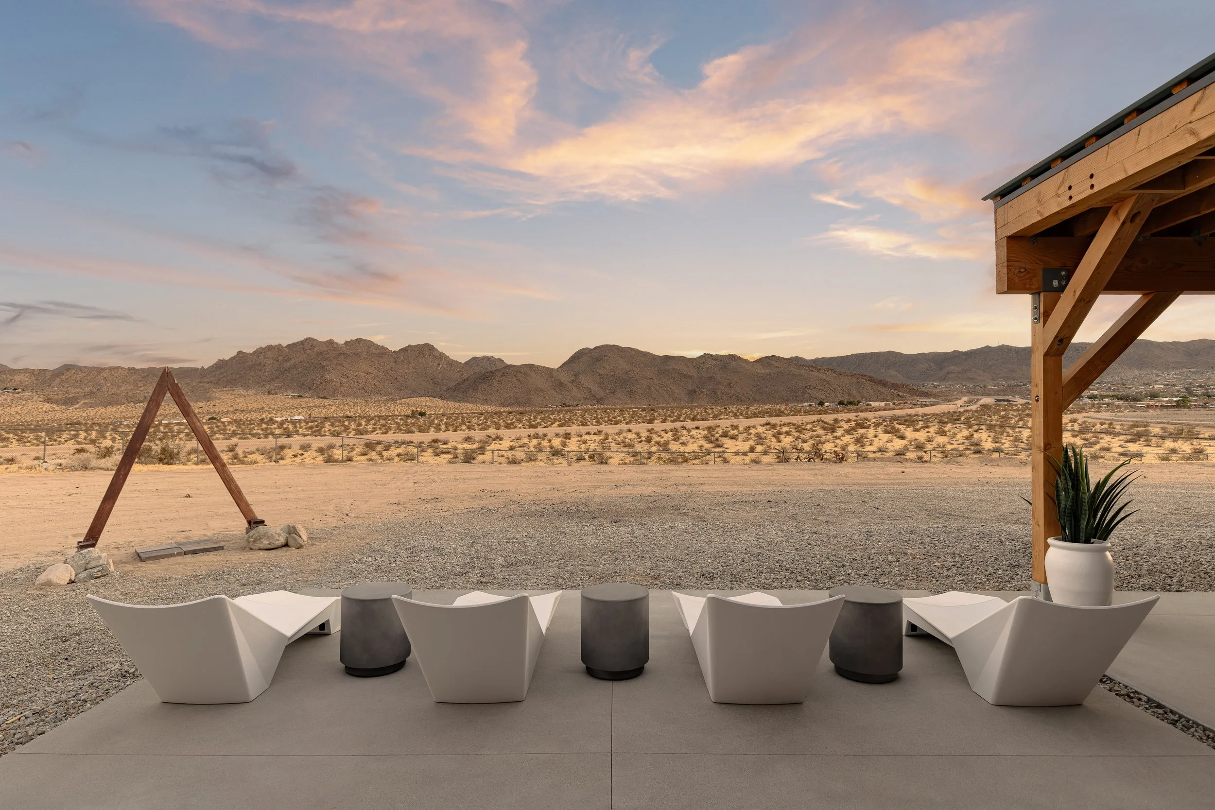 Outdoor seating area with six white geometric chairs and three black stools on a patio, overlooking a desert landscape with mountains in the distance under a colorful sky at sunset