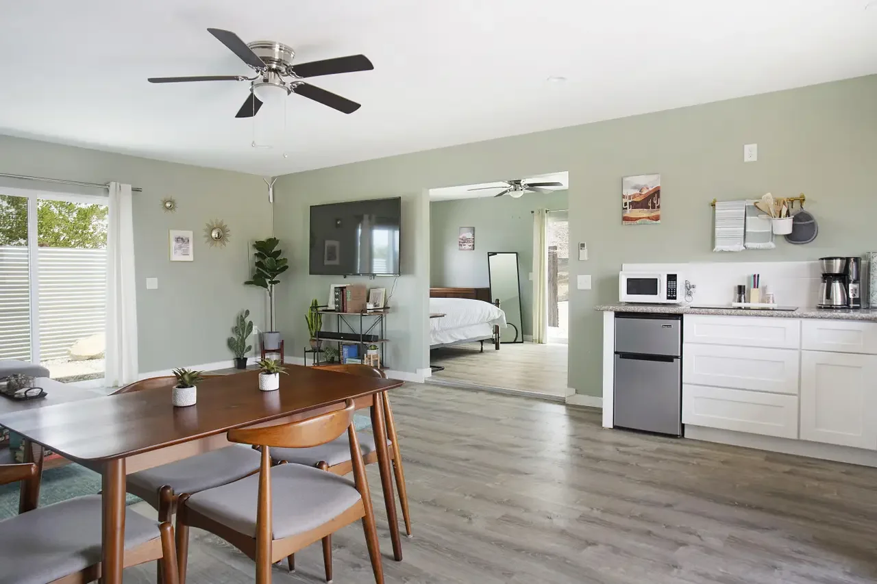 Open-concept living and dining area with light green walls, a ceiling fan, and wood flooring. The dining table has chairs and small potted plants. The living area includes wall decor, a TV, and potted plants. A doorway leads to a bedroom with a bed a