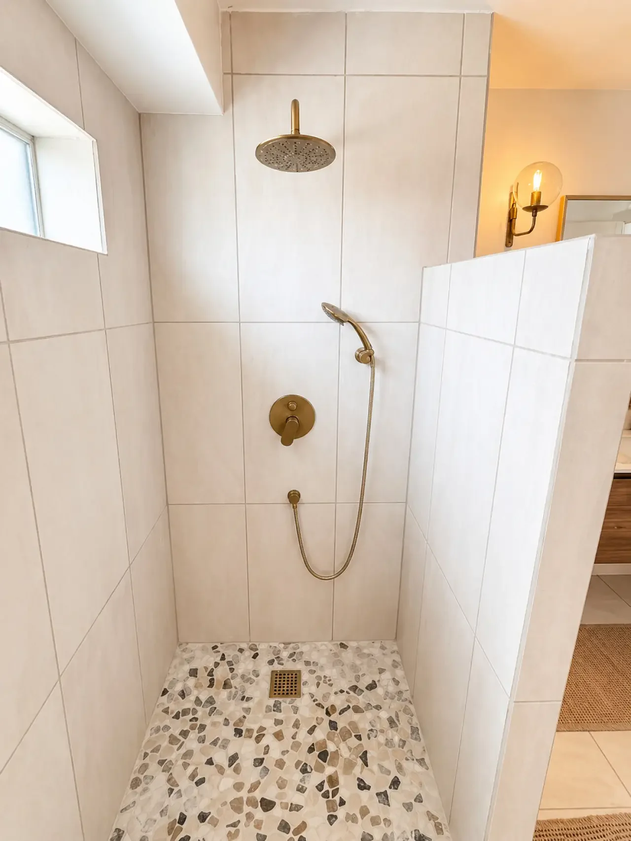 A modern shower with a rain showerhead, a handheld shower wand, white tiled walls, a small window near the ceiling, and a mosaic tile floor with a floor drain.