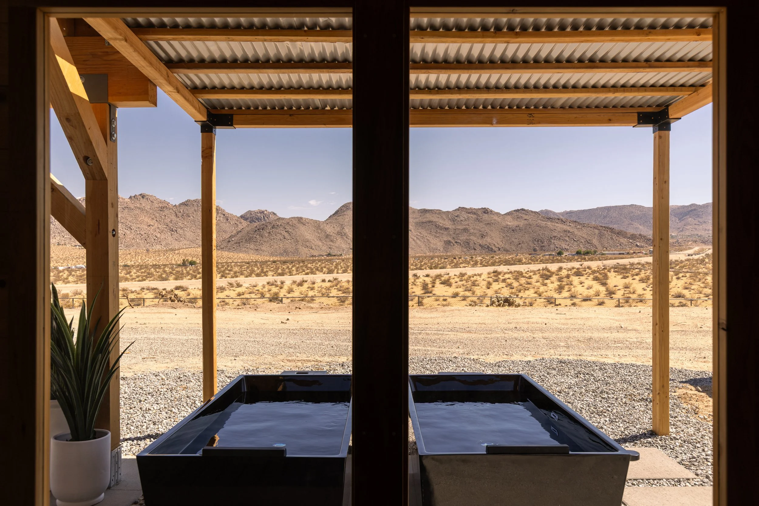 View from inside a structure looking out at a desert landscape with mountains in the distance. The structure has wooden posts, a corrugated metal roof, and a black hot tub or pond in the foreground.
