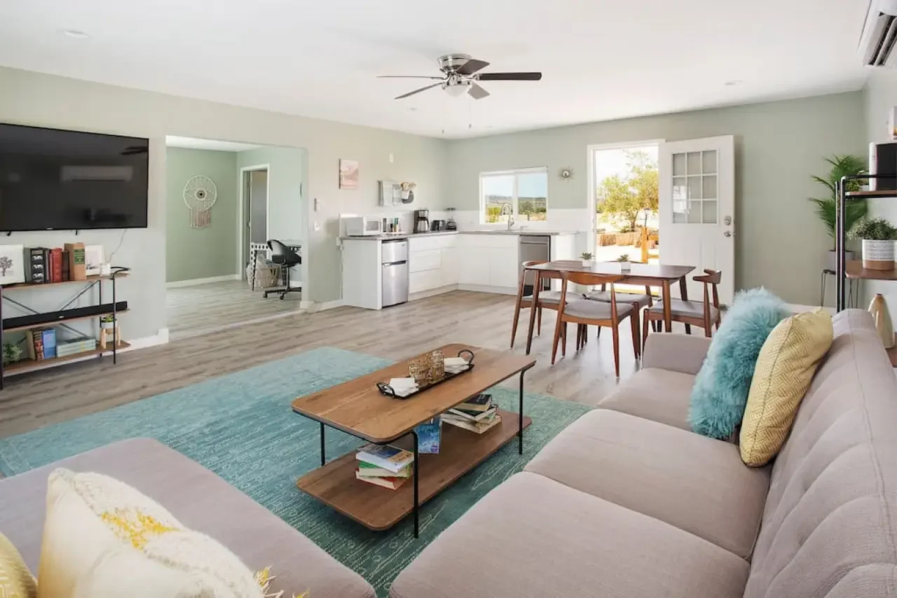 Open-plan living room and kitchen with a beige sofa, wooden coffee table, dining table with chairs, and kitchen appliances, with a sliding door leading outside.