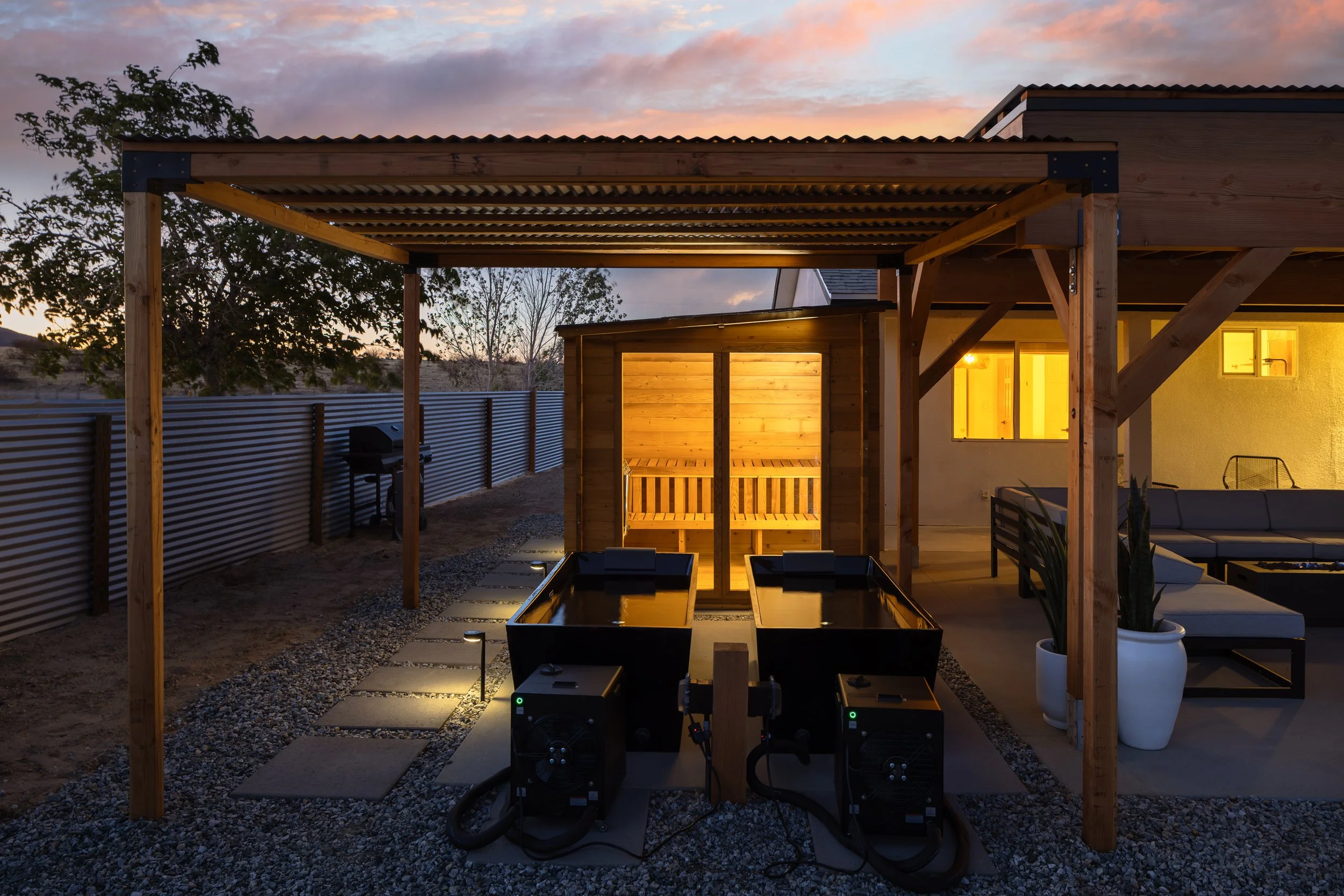A modern outdoor patio area during sunset with a wooden pergola structure, potted plants, a fence, and a seating area with furniture, illuminated by warm interior lights.