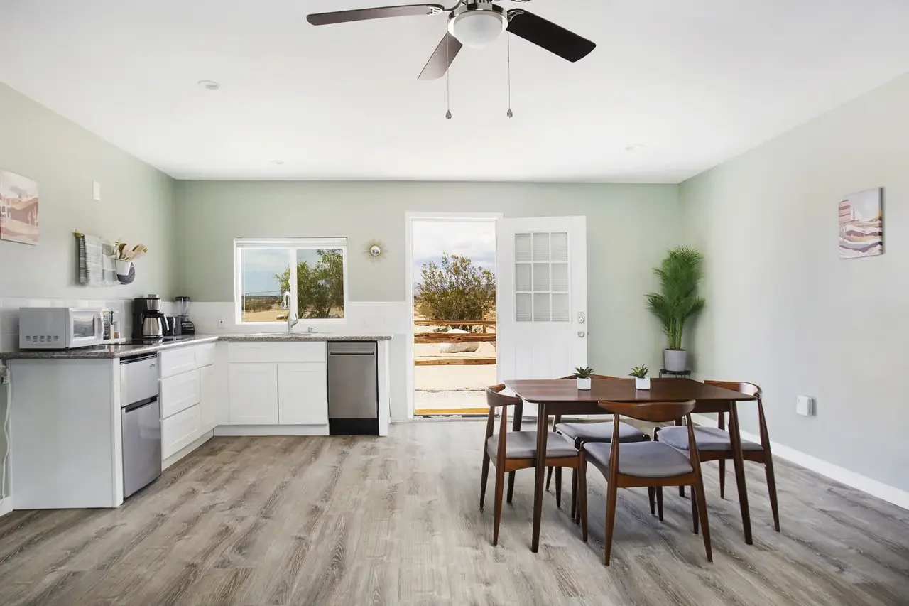 A modern kitchen and dining area with light green walls, white cabinets, a small dining table with six chairs, large window and door leading outside, wooden flooring, ceiling fan, and potted plants.