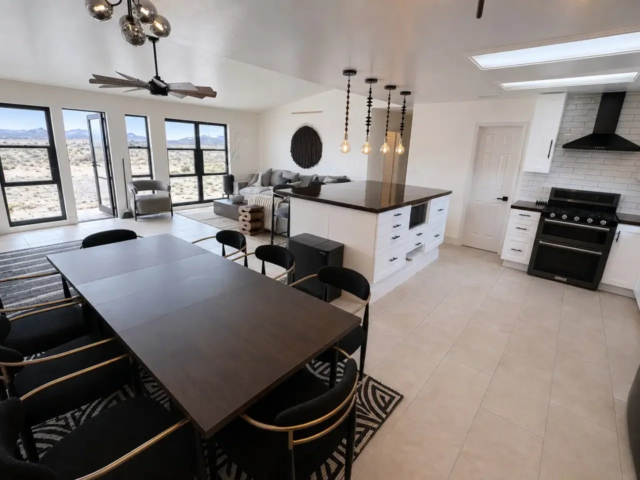 Open-concept kitchen and living room with large windows, dining table, black chairs, white cabinets, black stove, and a coffee table with a sectional sofa.