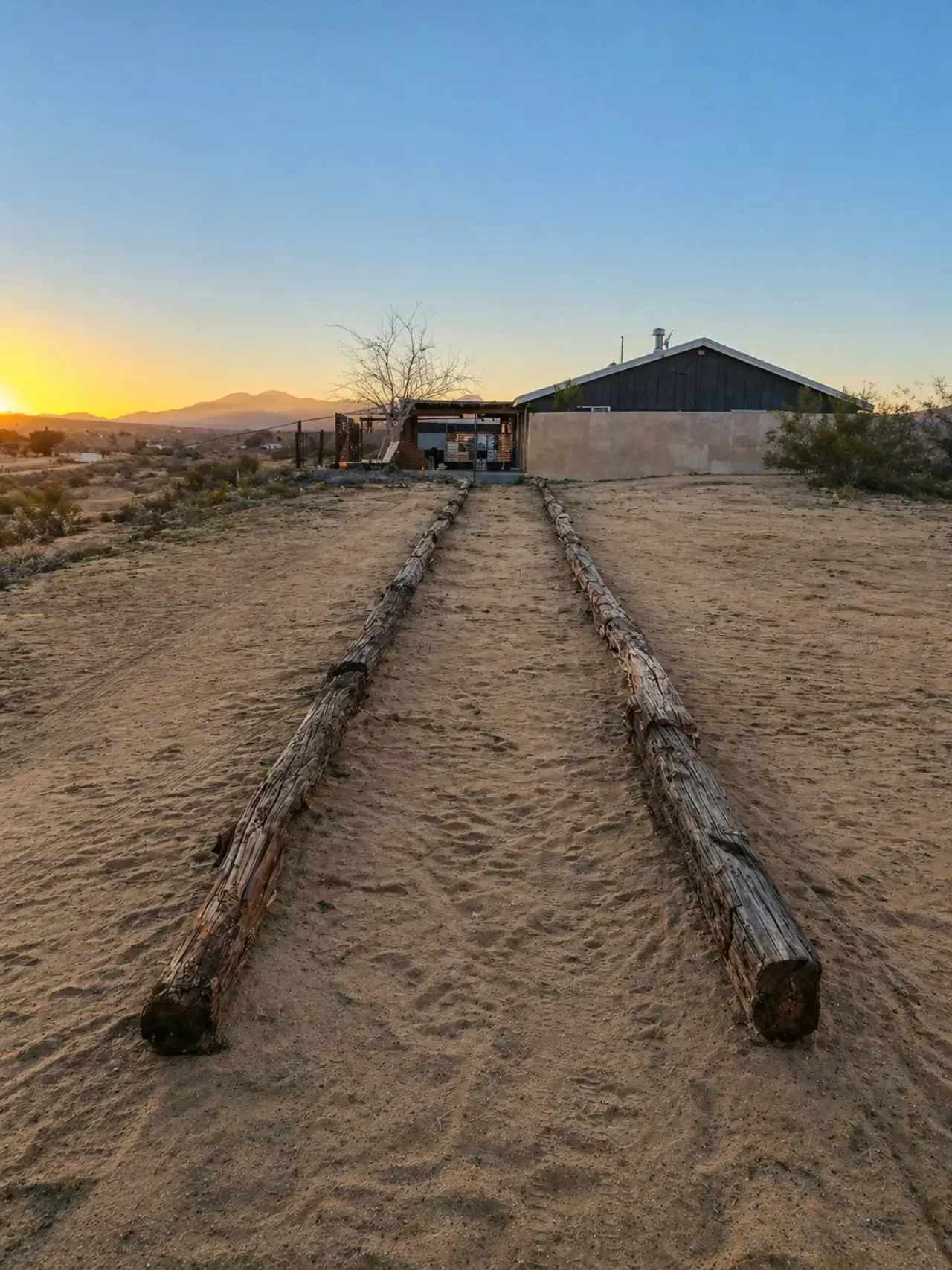 Desert landscape at sunset with a house in the distance, a large dead tree, and wooden railroad ties leading to the house.