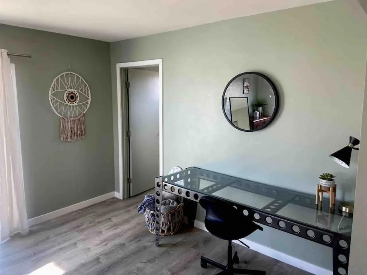 A home office with pale green walls, a glass-top desk with circular cutouts, a black office chair, a wall mirror, a woven wall hanging, and a laundry basket filled with clothes.