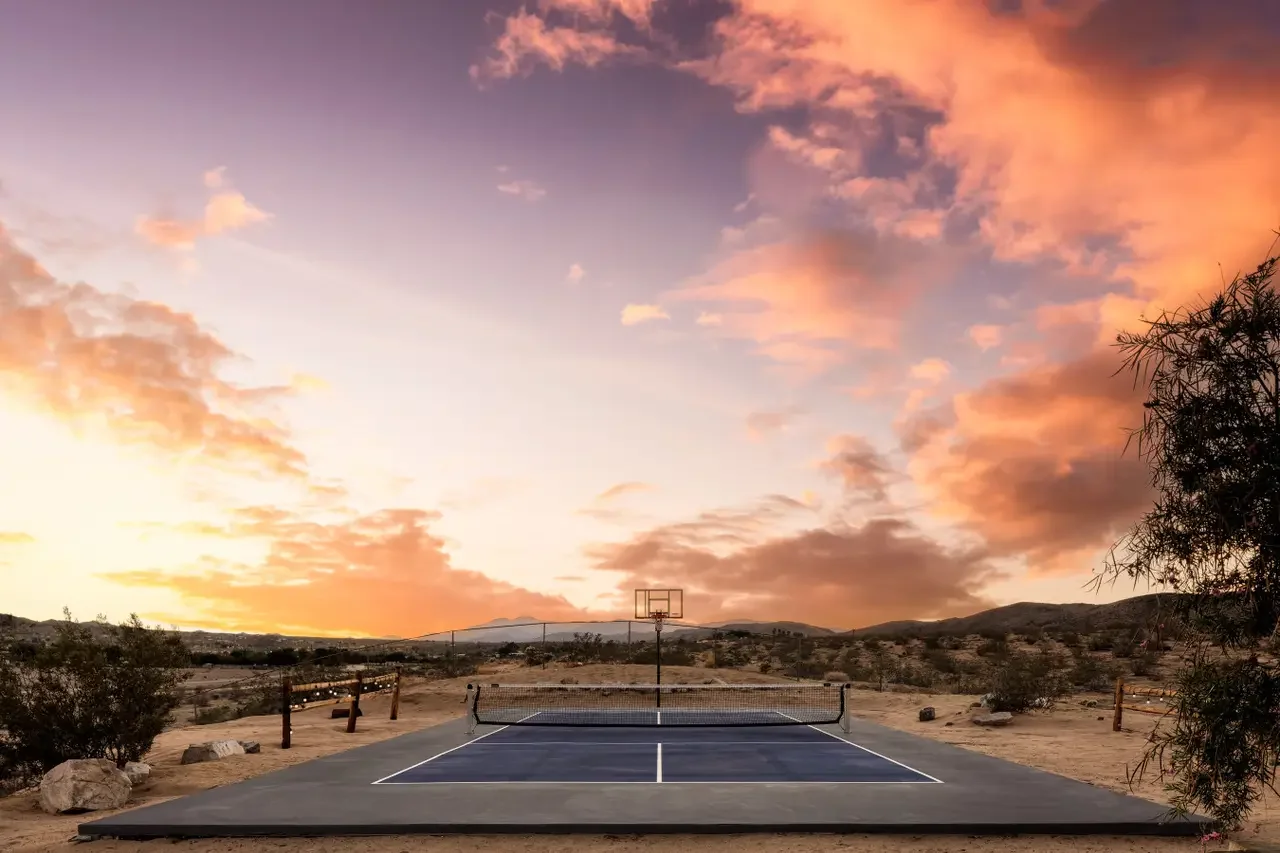 An outdoor tennis court in a desert landscape at sunset with pink and orange clouds, surrounded by sparse vegetation and distant hills.