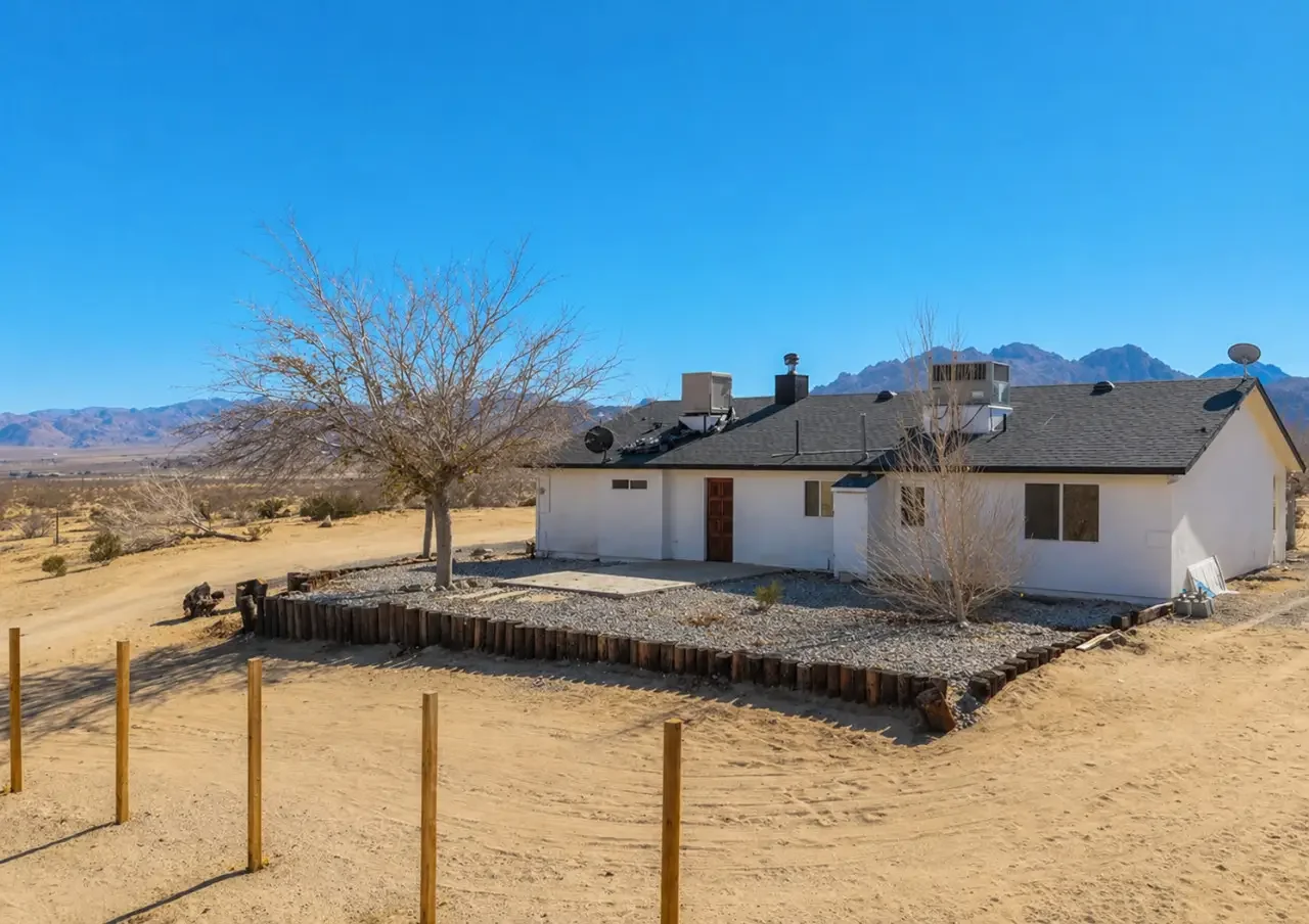 Single-story white house with black roof and dry desert landscape, fenced yard with gravel decoration, leafless trees, mountains in the background.