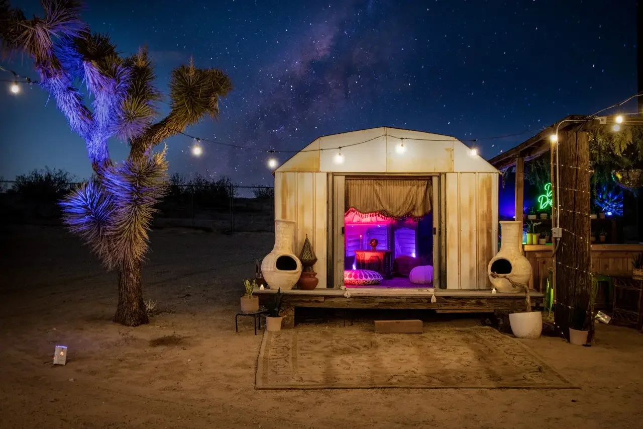 Night scene of a desert with a small wooden cabin lit by string lights, with a starry sky and the Milky Way visible. There is a joshua tree on the left decorated with purple lights, and pottery details near the cabin entrance.