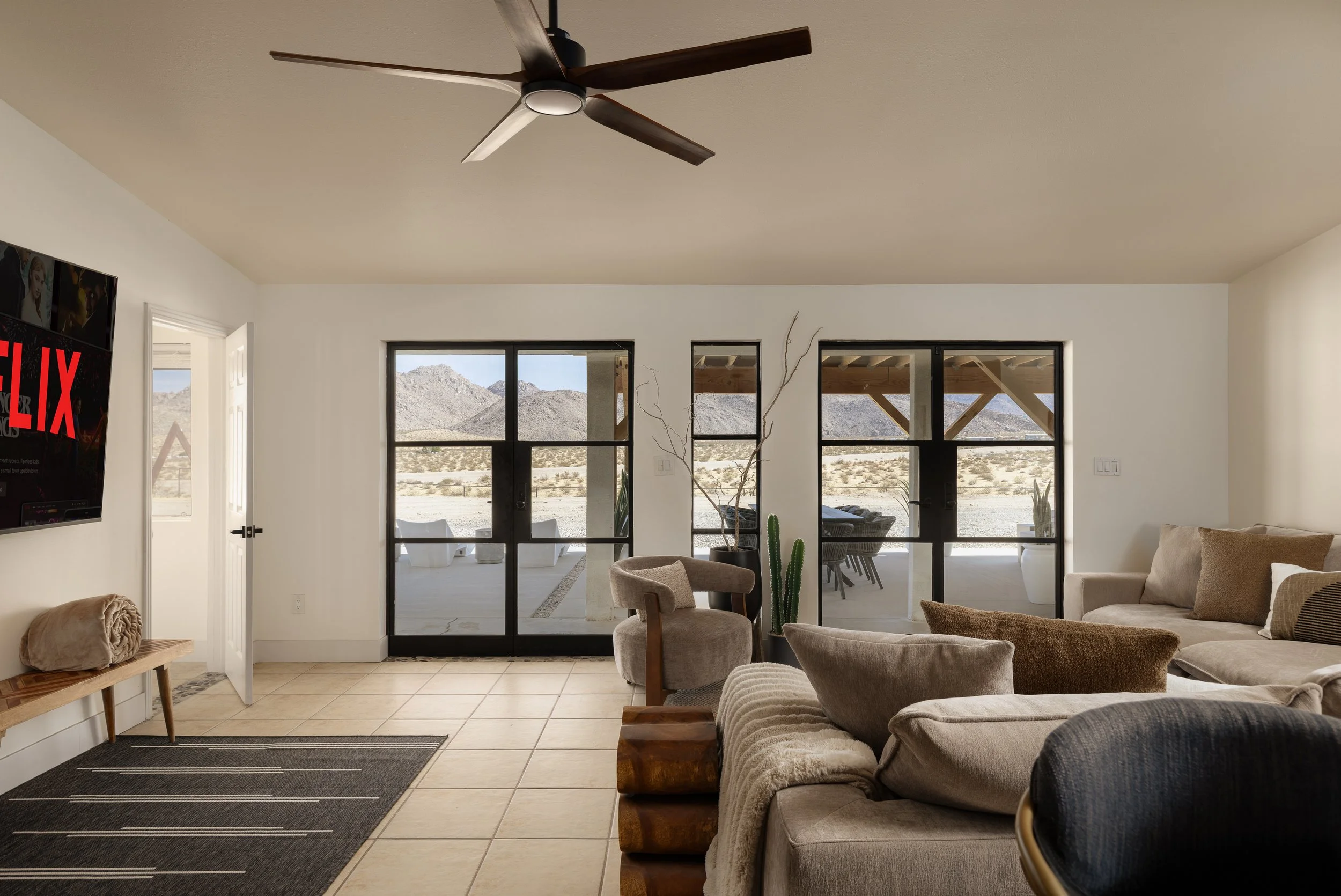 Living room with beige tiled floor, beige sofas, a black sliding glass door leading to a desert landscape with mountains, and a ceiling fan with dark wooden blades.