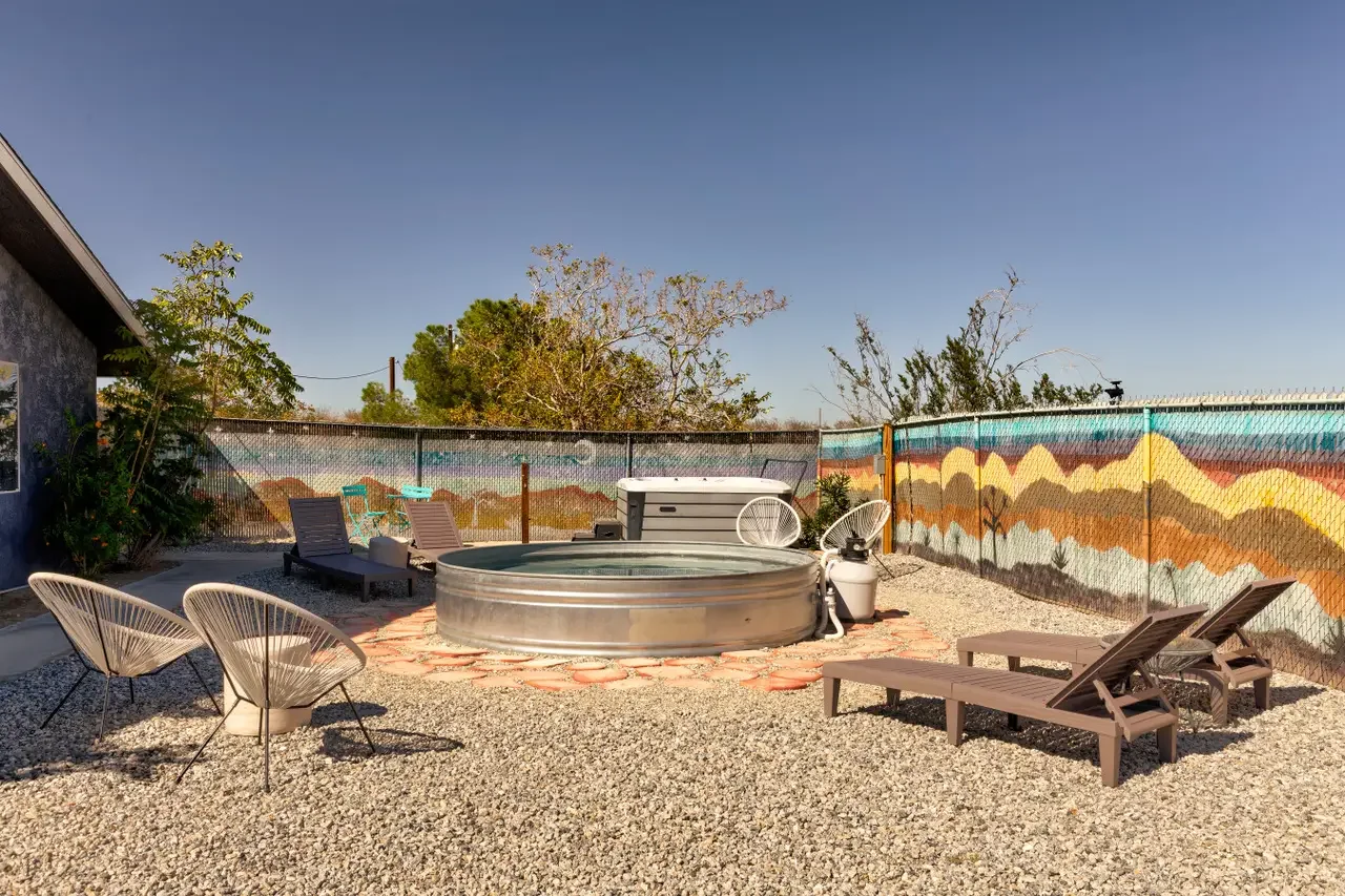 A backyard with a hot tub, lounge chairs, and a colorful mountain mural fence under a clear blue sky.