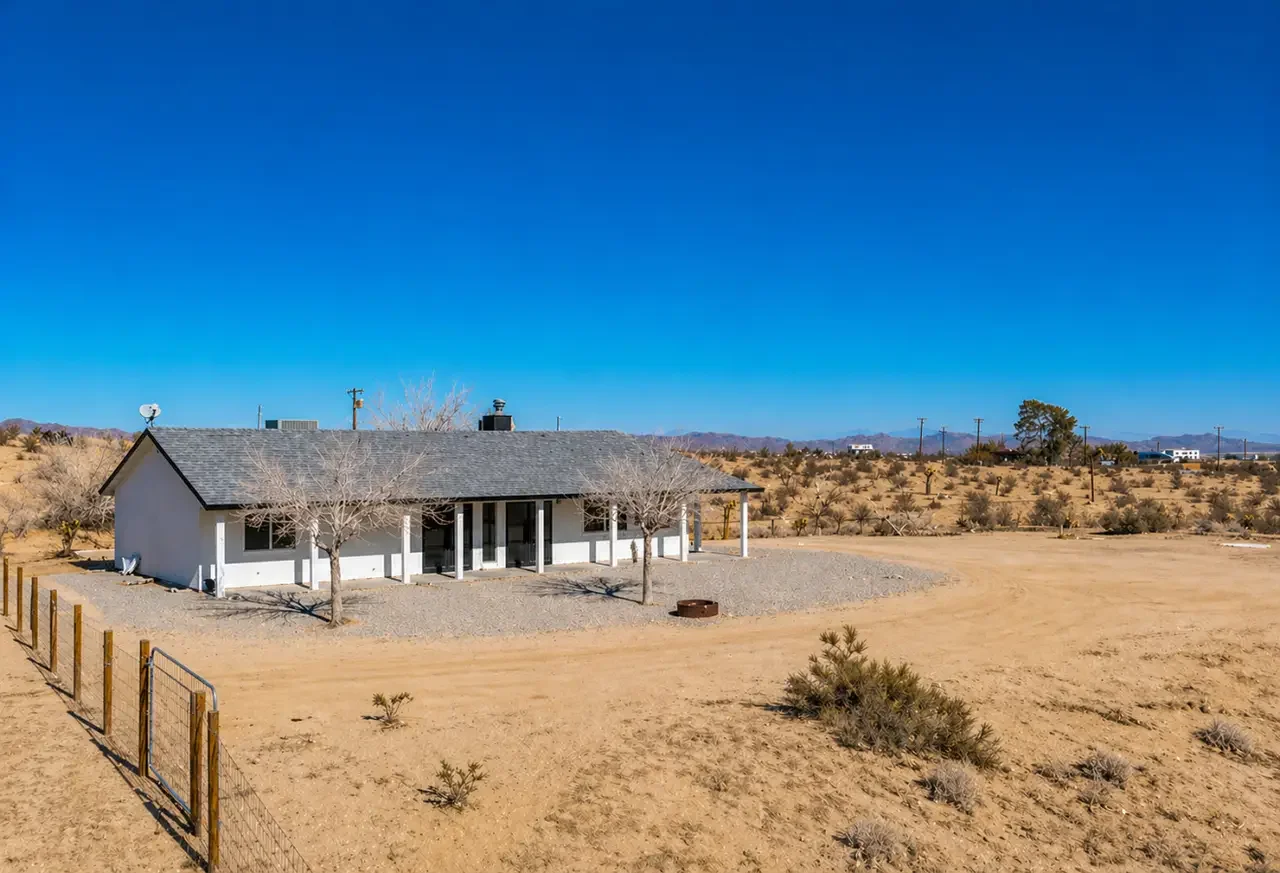 A single-story house with white walls and a gray roof in a desert landscape under a clear blue sky. The house has a porch, leafless trees in front, and a fenced yard.