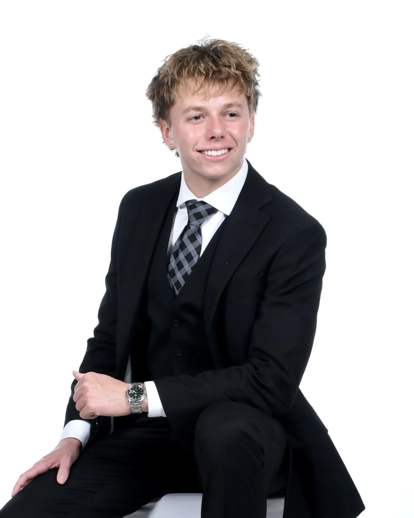 Young man in a black suit, white shirt, and patterned tie sitting with a smile, against a white background.