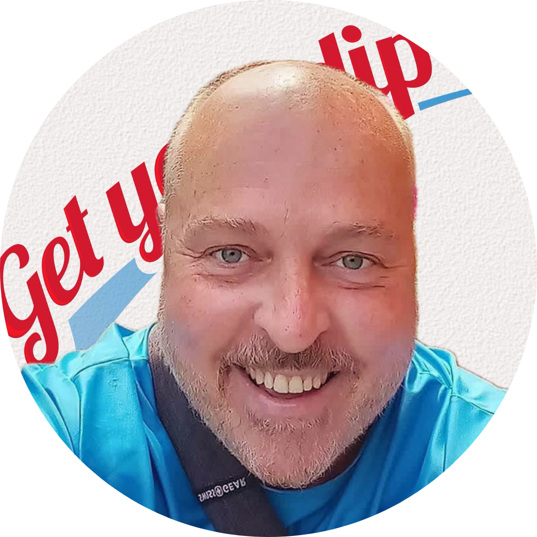 A smiling man with a beard and blue eyes taking a selfie in front of a sign that says 'Get y' with a red arrow pointing to the right.